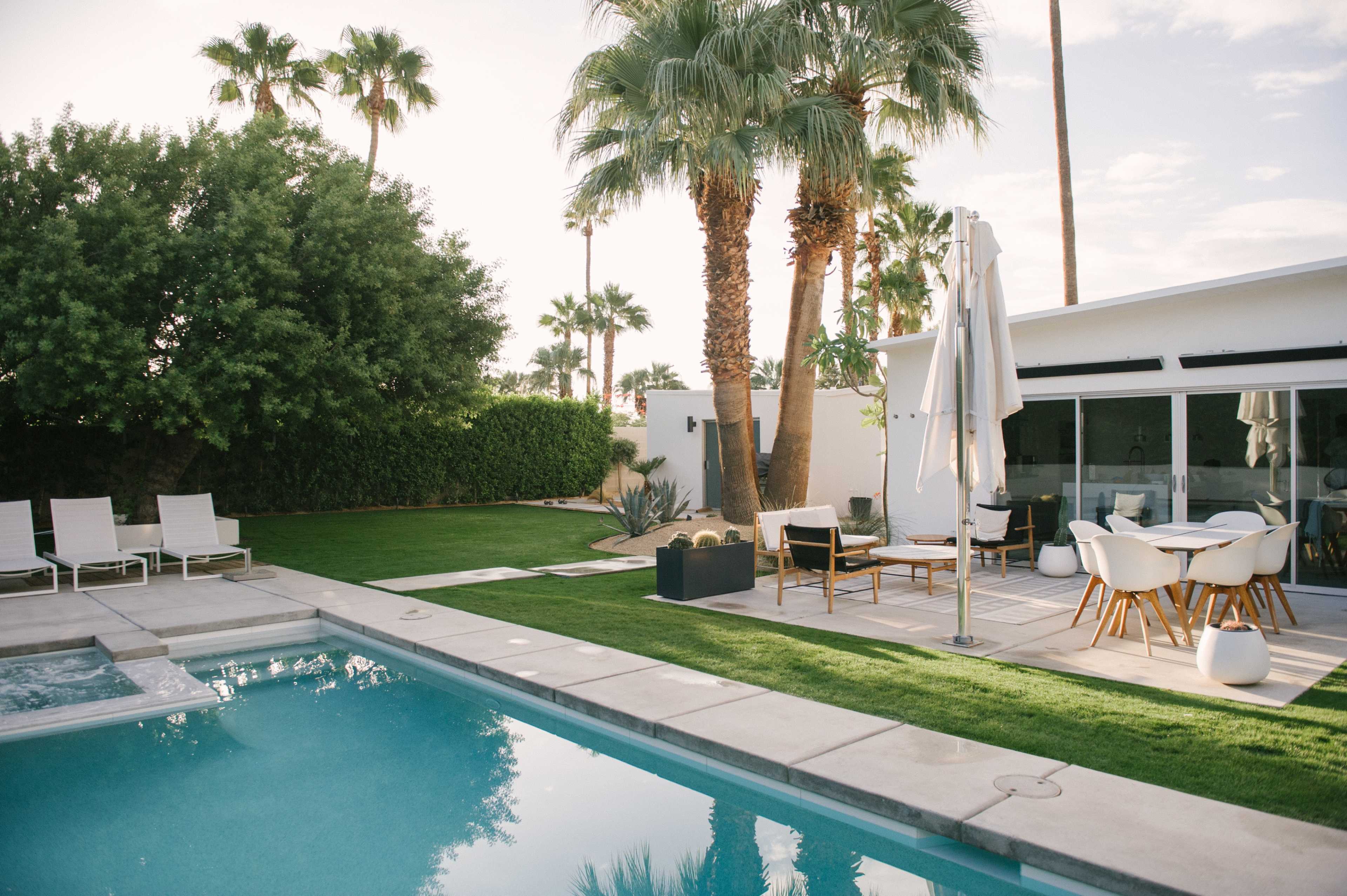 A landscaped backyard features a swimming pool, lounge chairs, a dining area, and palm trees under a clear sky.