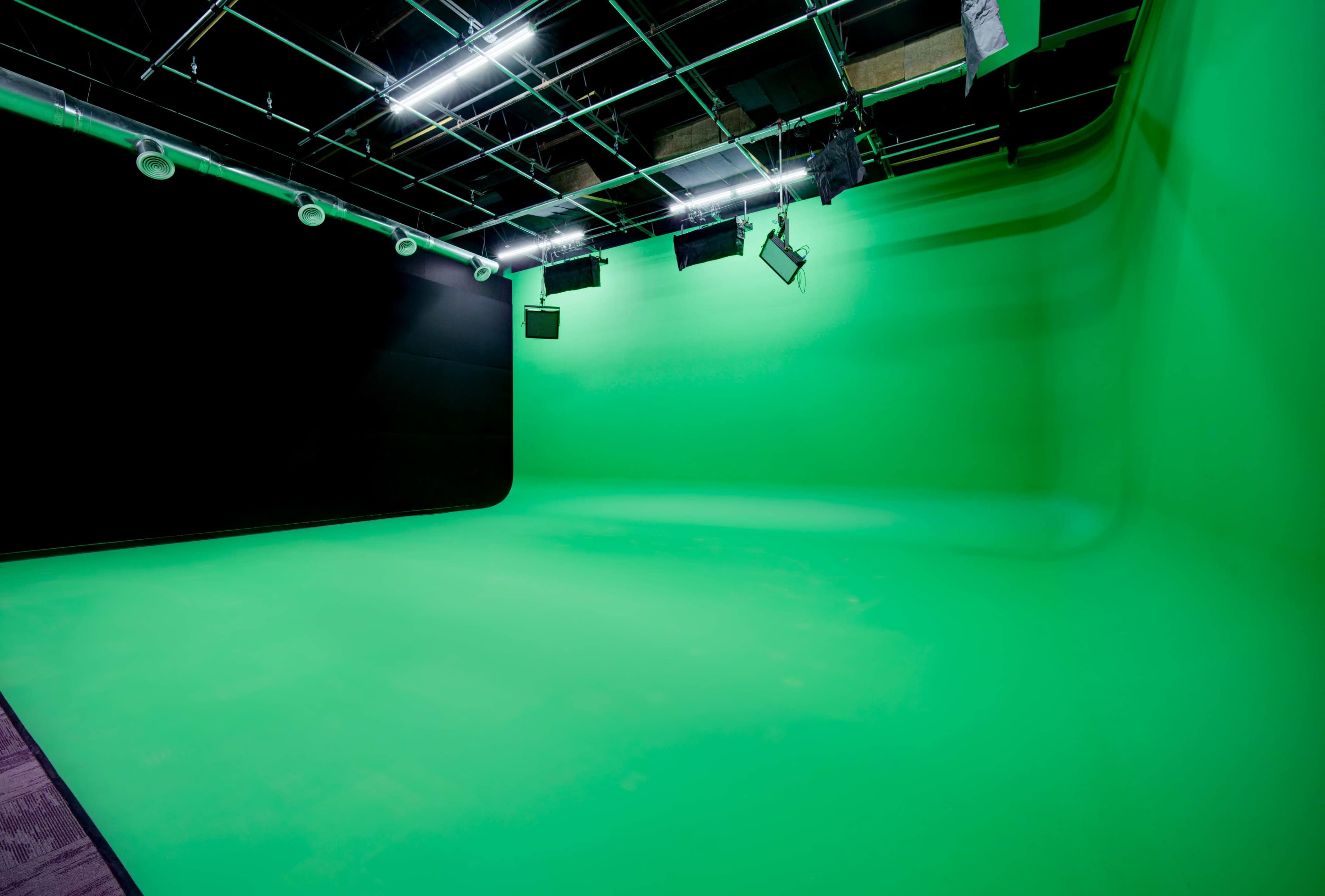 The image shows a large green screen studio with a seamless green backdrop and overhead lighting fixtures.