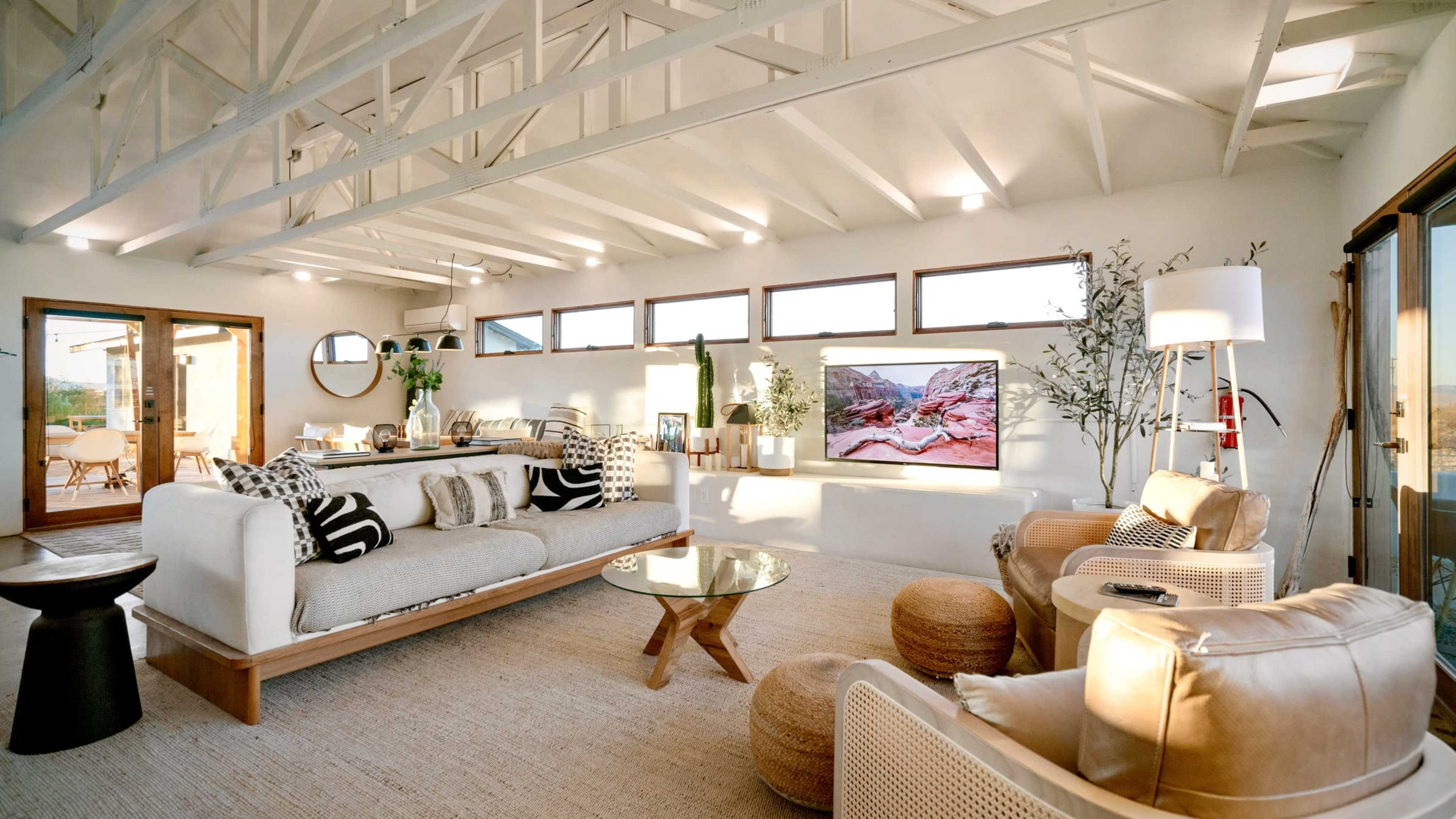 The image shows a modern living room with high, white-beamed ceilings, a large sofa, a coffee table, and a wall-mounted television.