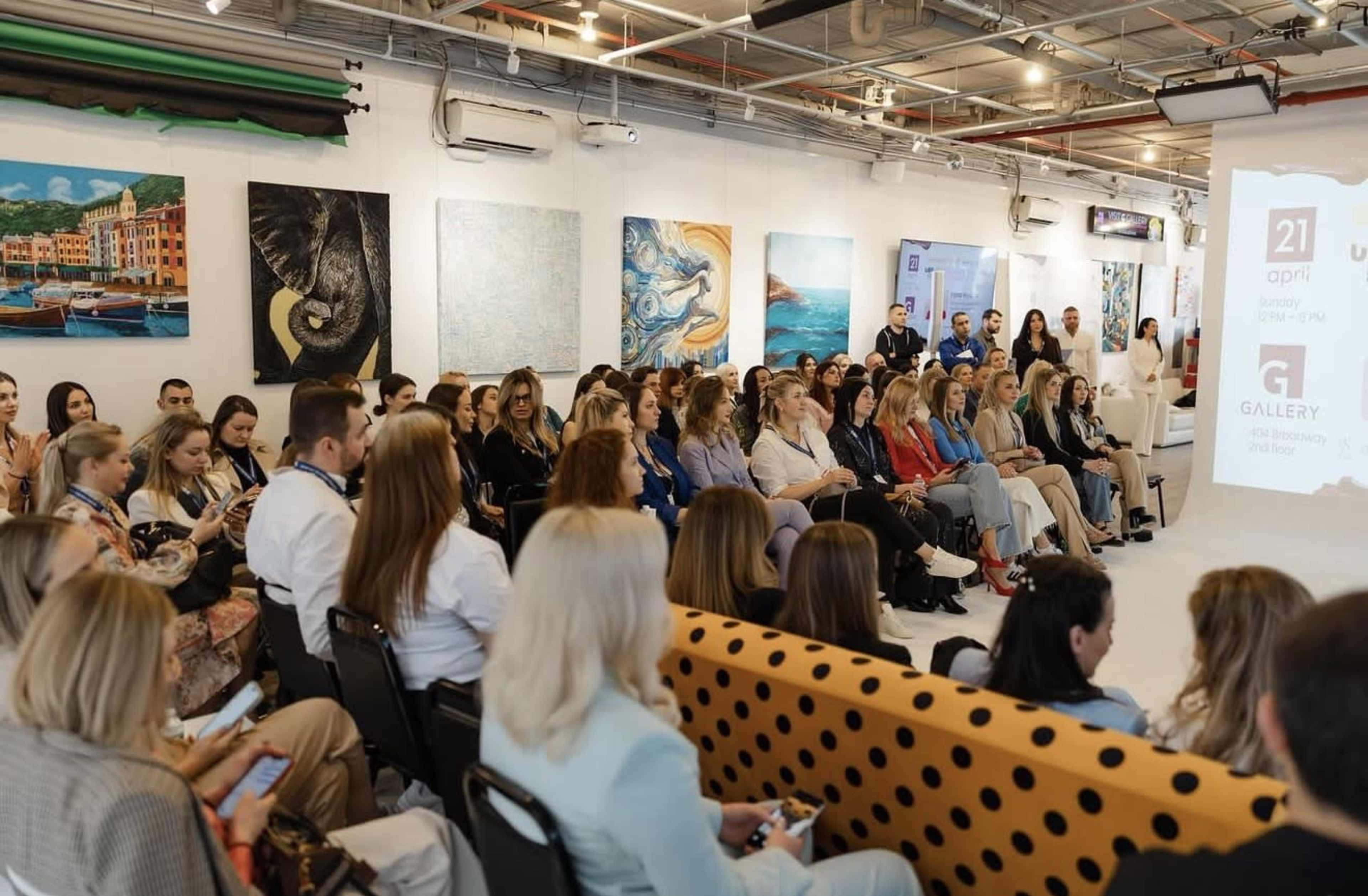 A large audience of people is seated in a modern gallery space, surrounded by various colorful artworks displayed on the walls.