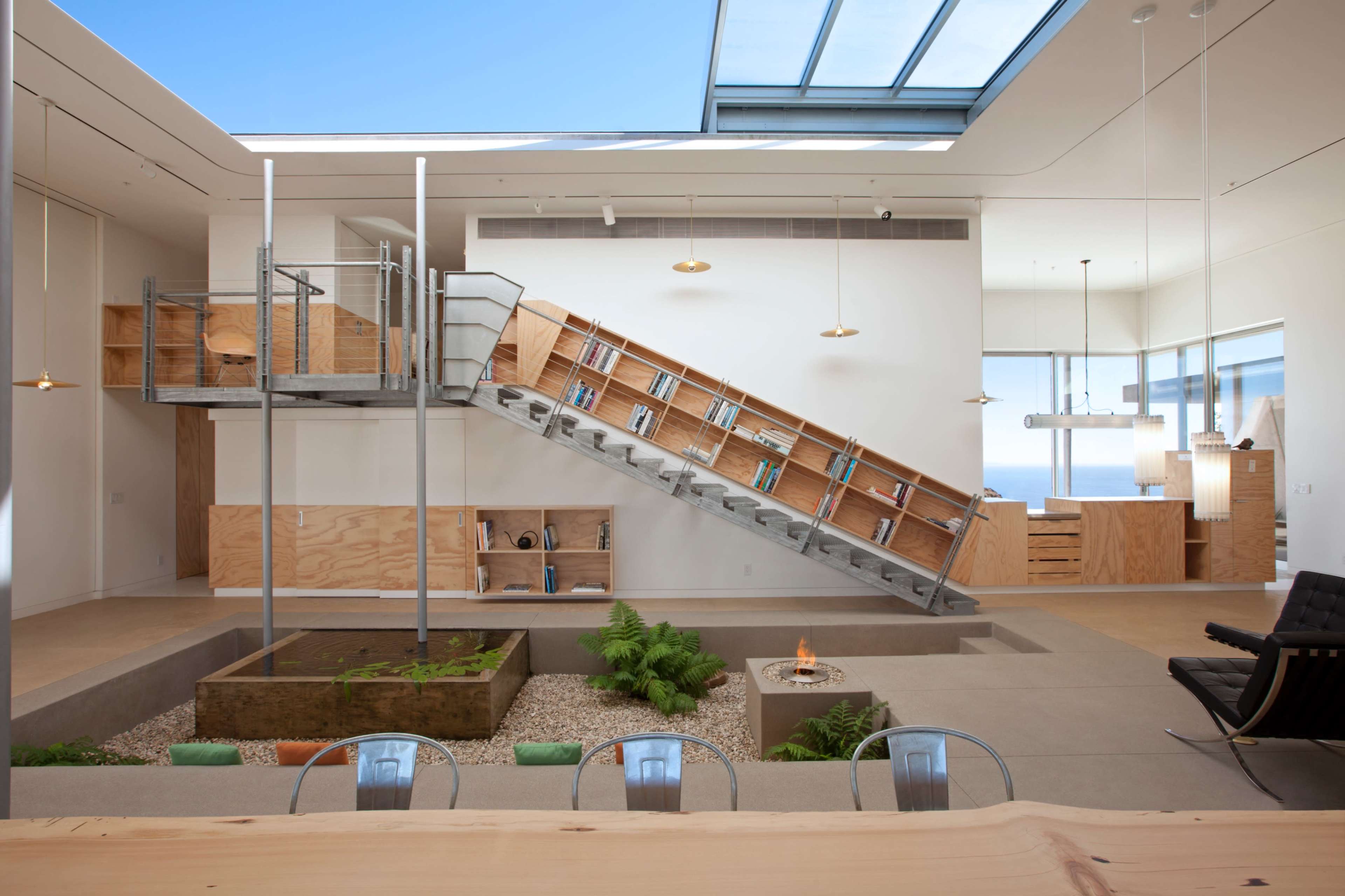 The image shows a modern interior space featuring an inclined bookshelf staircase, a central garden area, and large windows that allow natural light to flood in.