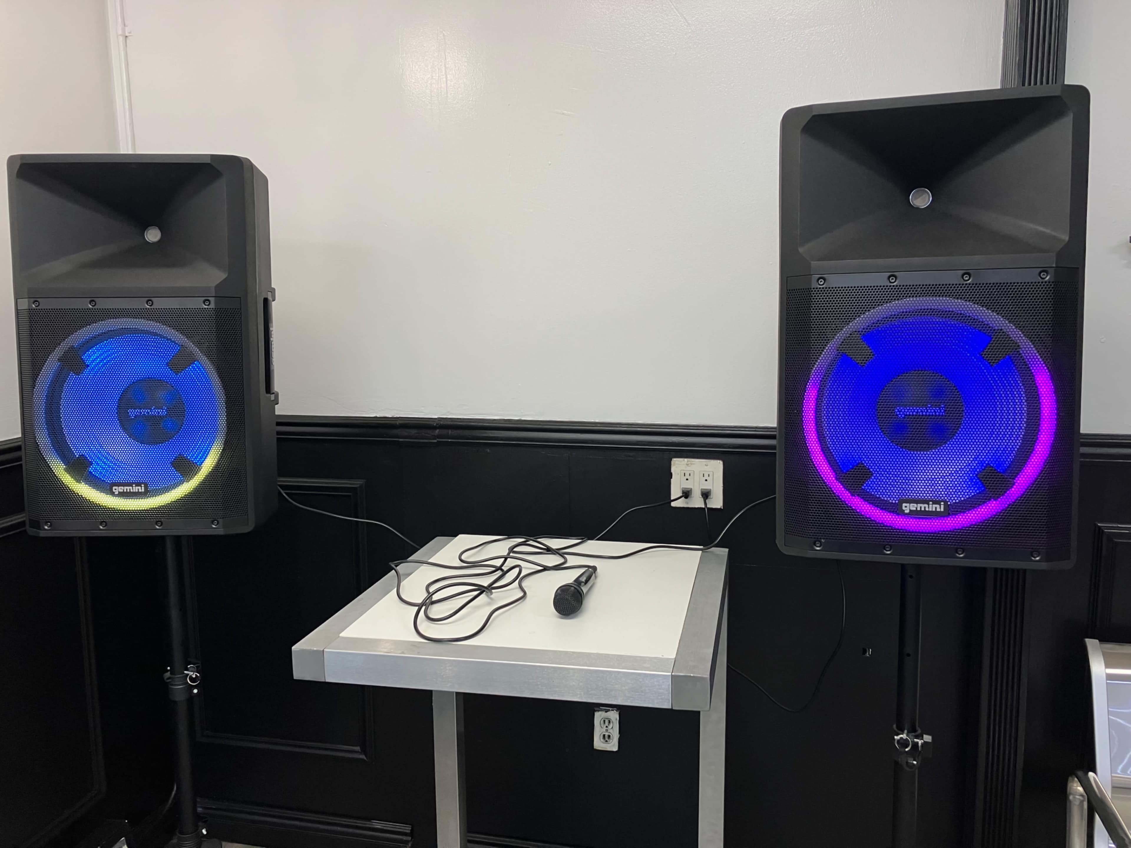 Two portable speakers with colorful LED lights are placed on stands next to a table in a room with black and white walls.