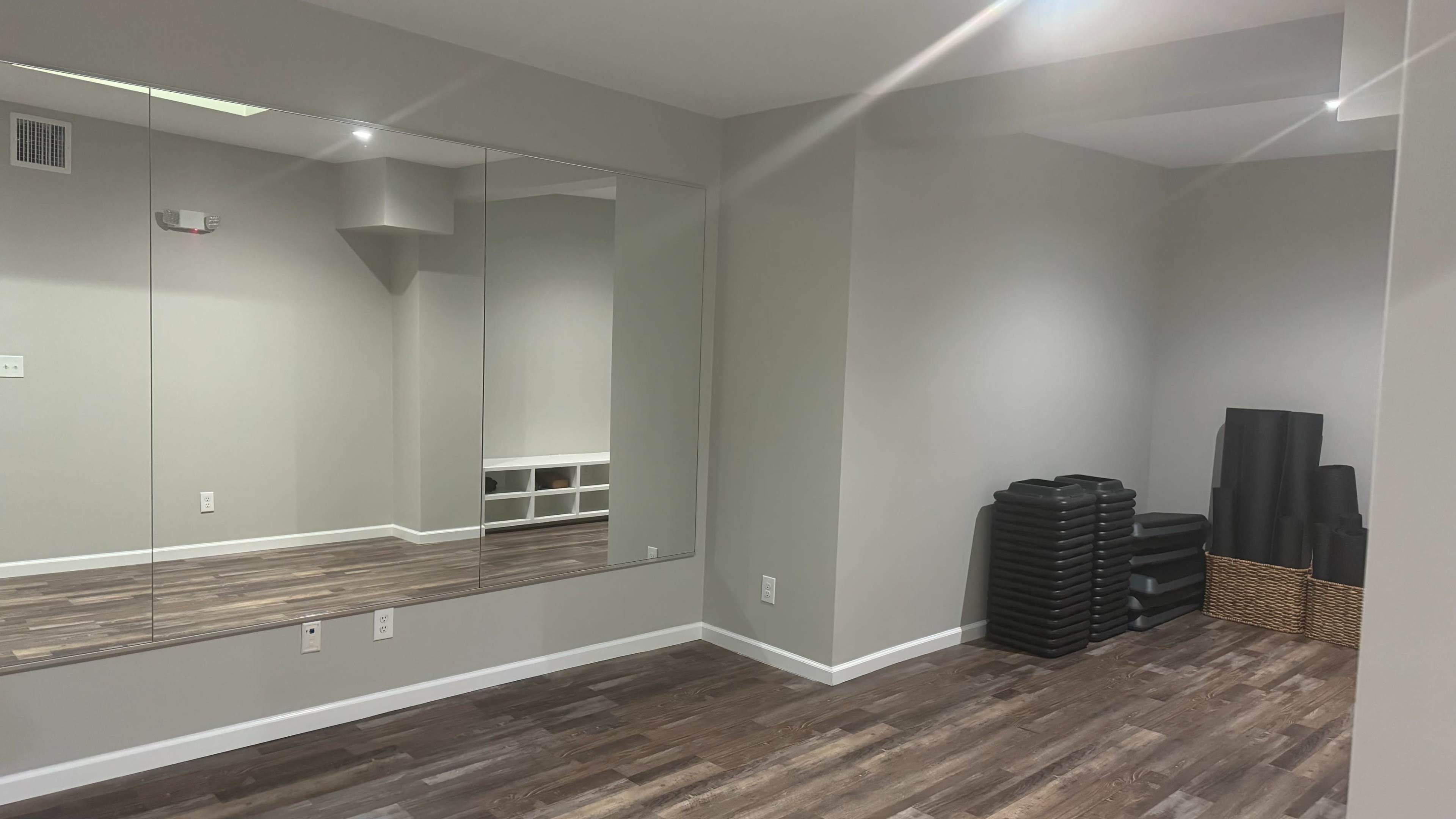 The image shows a light gray room with a large mirror on one wall, wooden flooring, and storage items, including black exercise mats and a wicker basket.