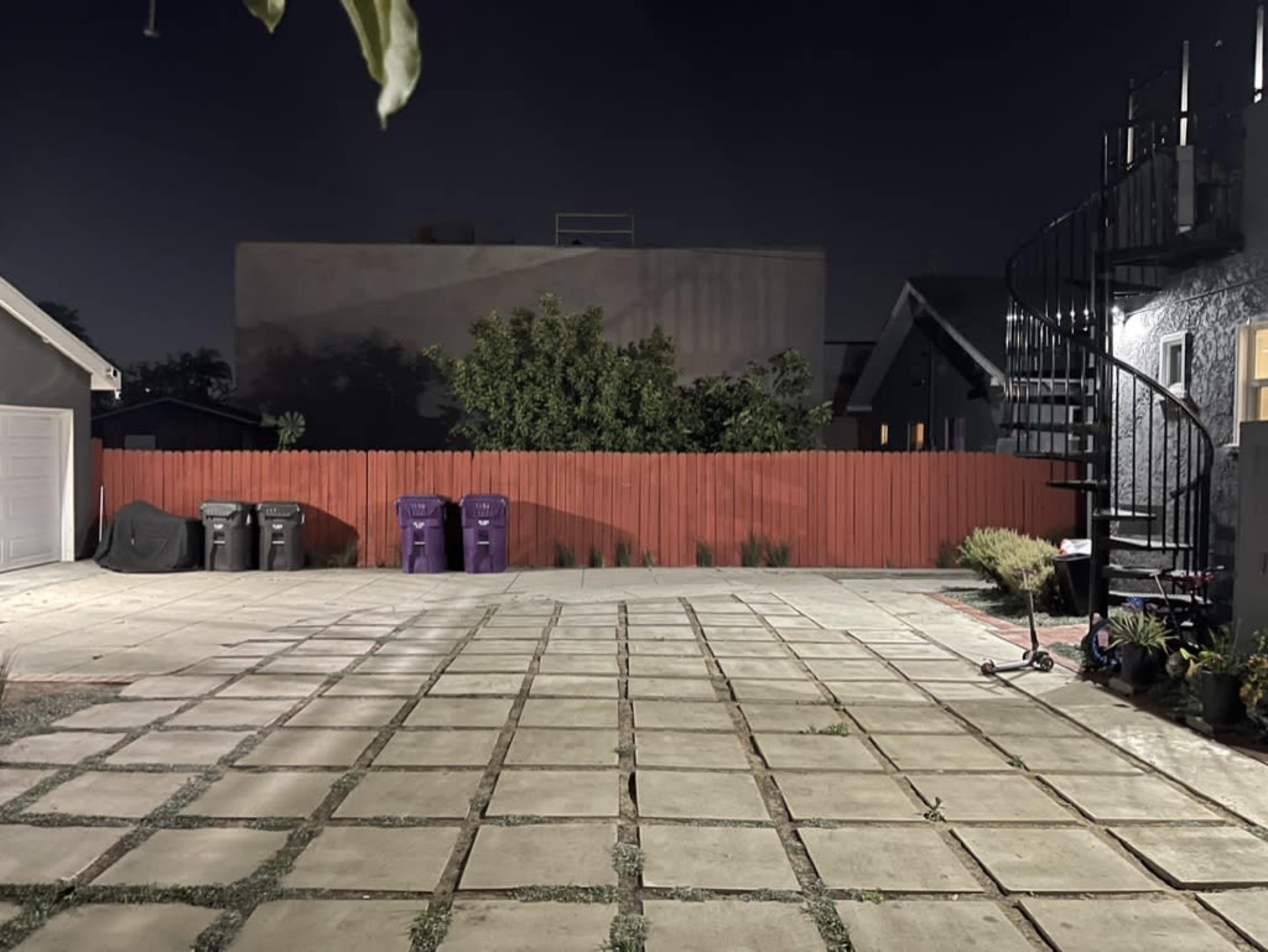 The image shows a spacious backyard at night, featuring a red fenced area, a spiral staircase, and several garbage bins along the side.