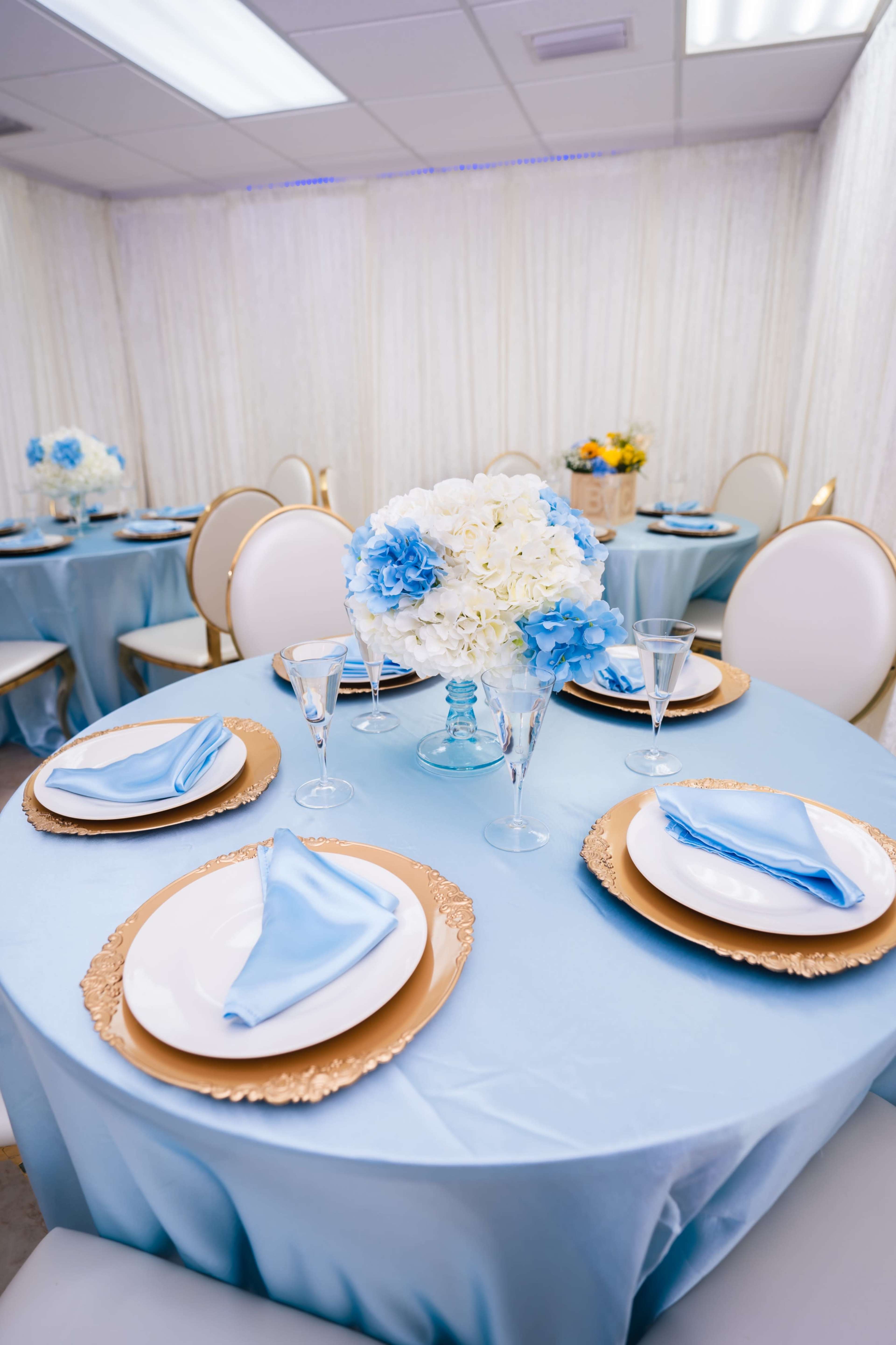 A neatly arranged banquet setup features round tables with blue tablecloths, white and blue floral centerpieces, and coordinated plates and napkins.