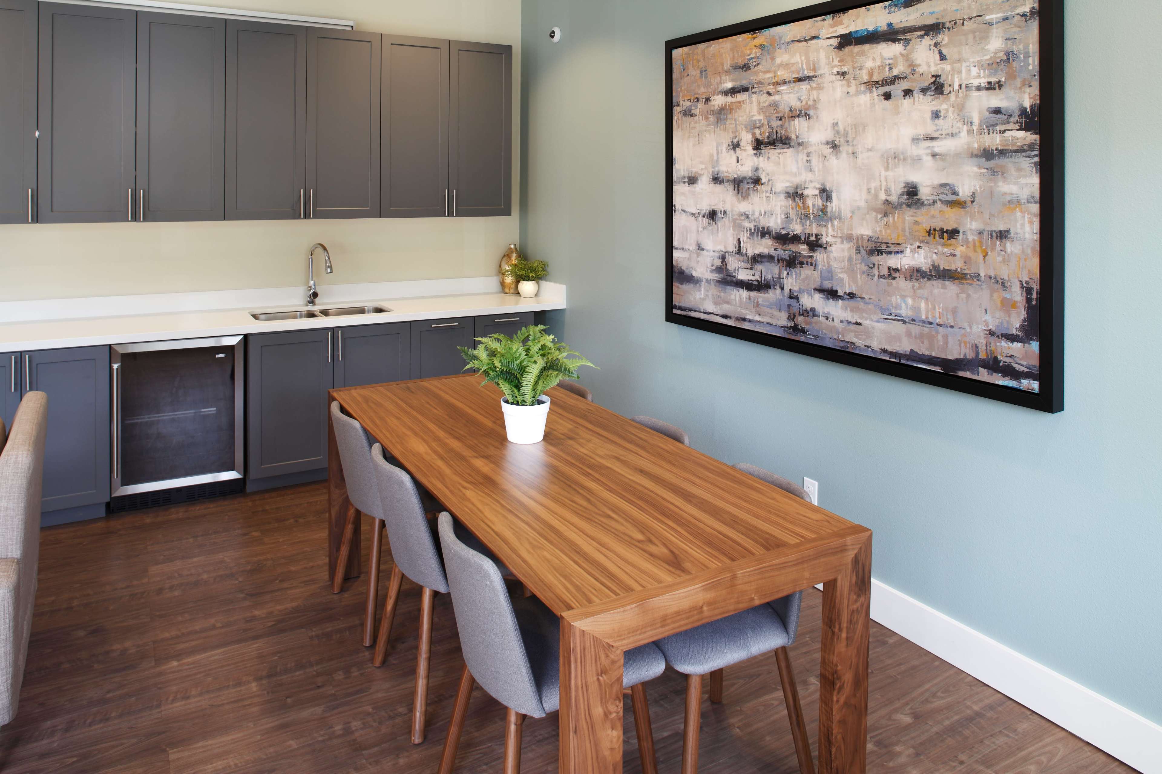 A modern dining area with a wooden table surrounded by gray chairs, a kitchen counter with cabinets, and a large abstract painting on the wall.