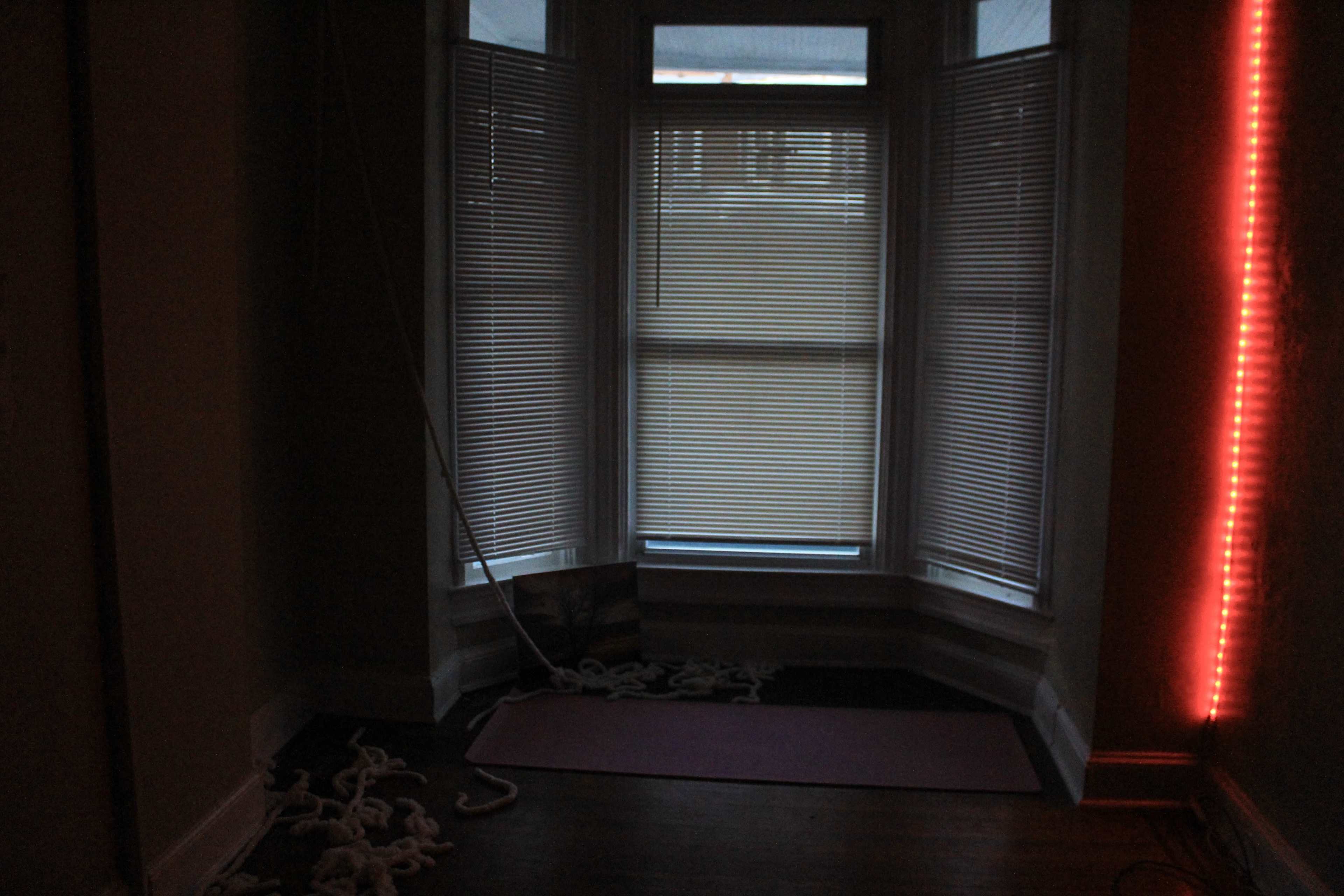 The image shows a dimly lit corner of a room with a window covered by blinds and a red light strip on the wall.