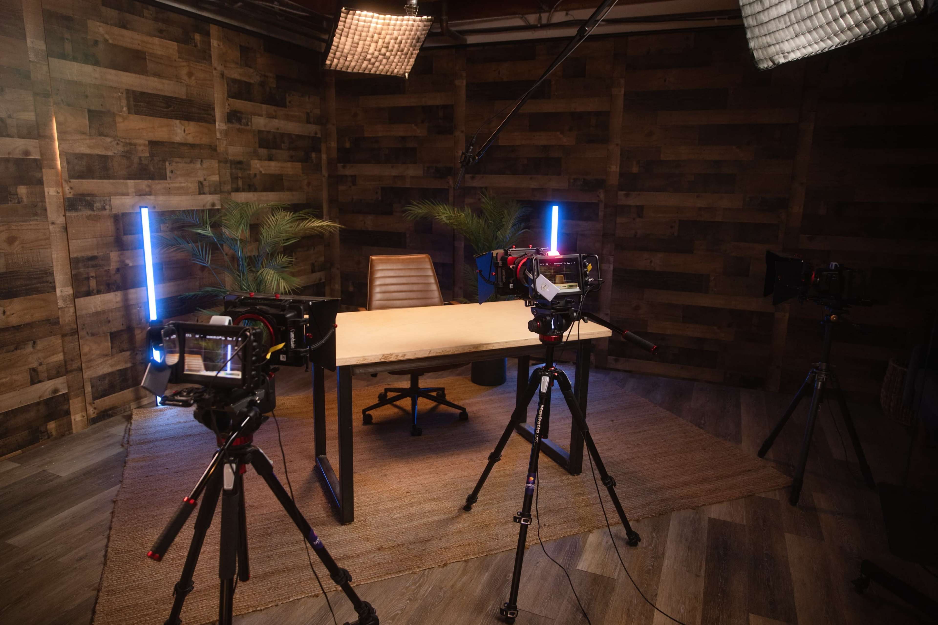 The image shows a photography studio with a wooden backdrop, a central table and chair, and professional camera equipment positioned on tripods.