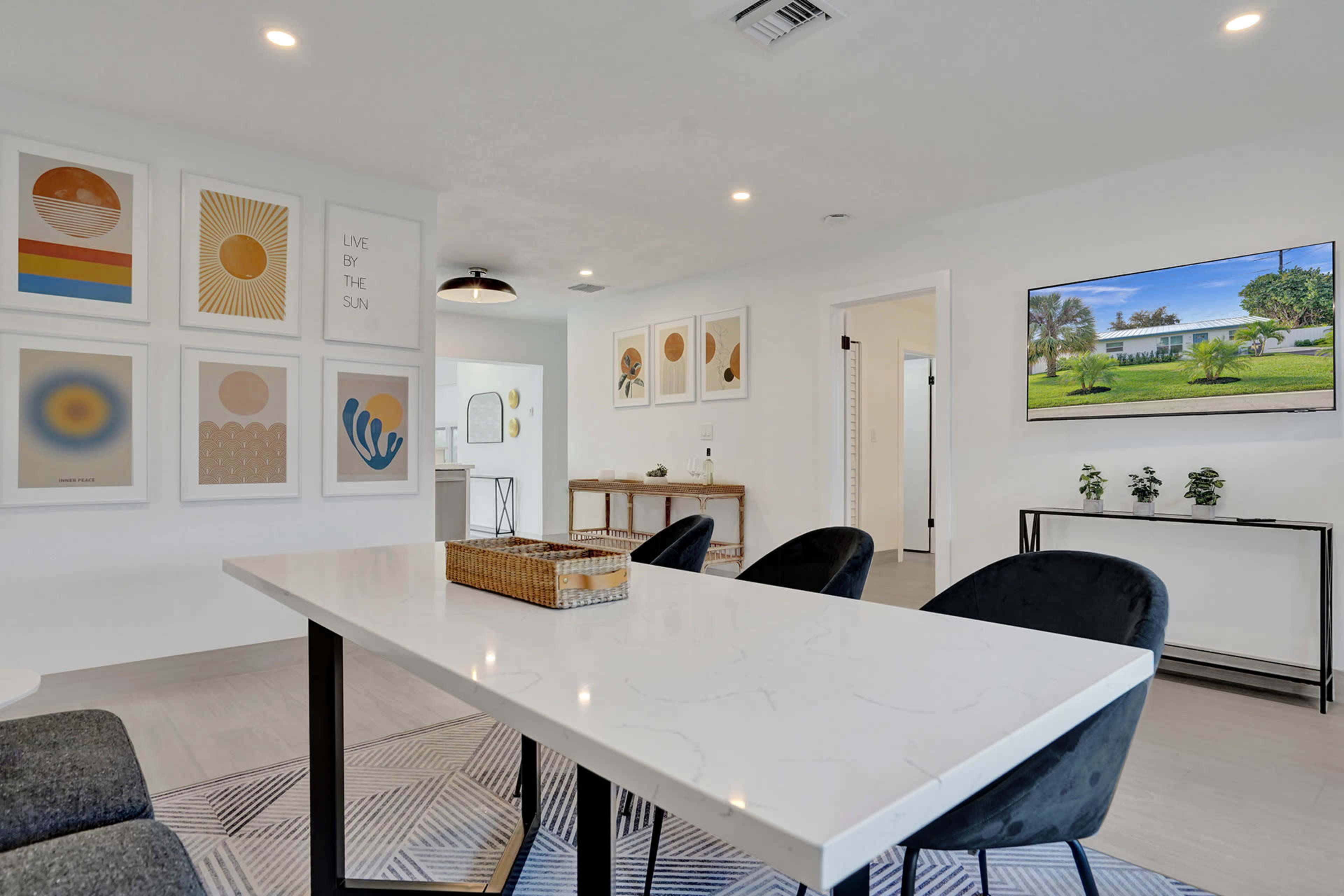 A modern dining area features a large table surrounded by black chairs, with framed artworks on the walls and a television displaying an outdoor scene.