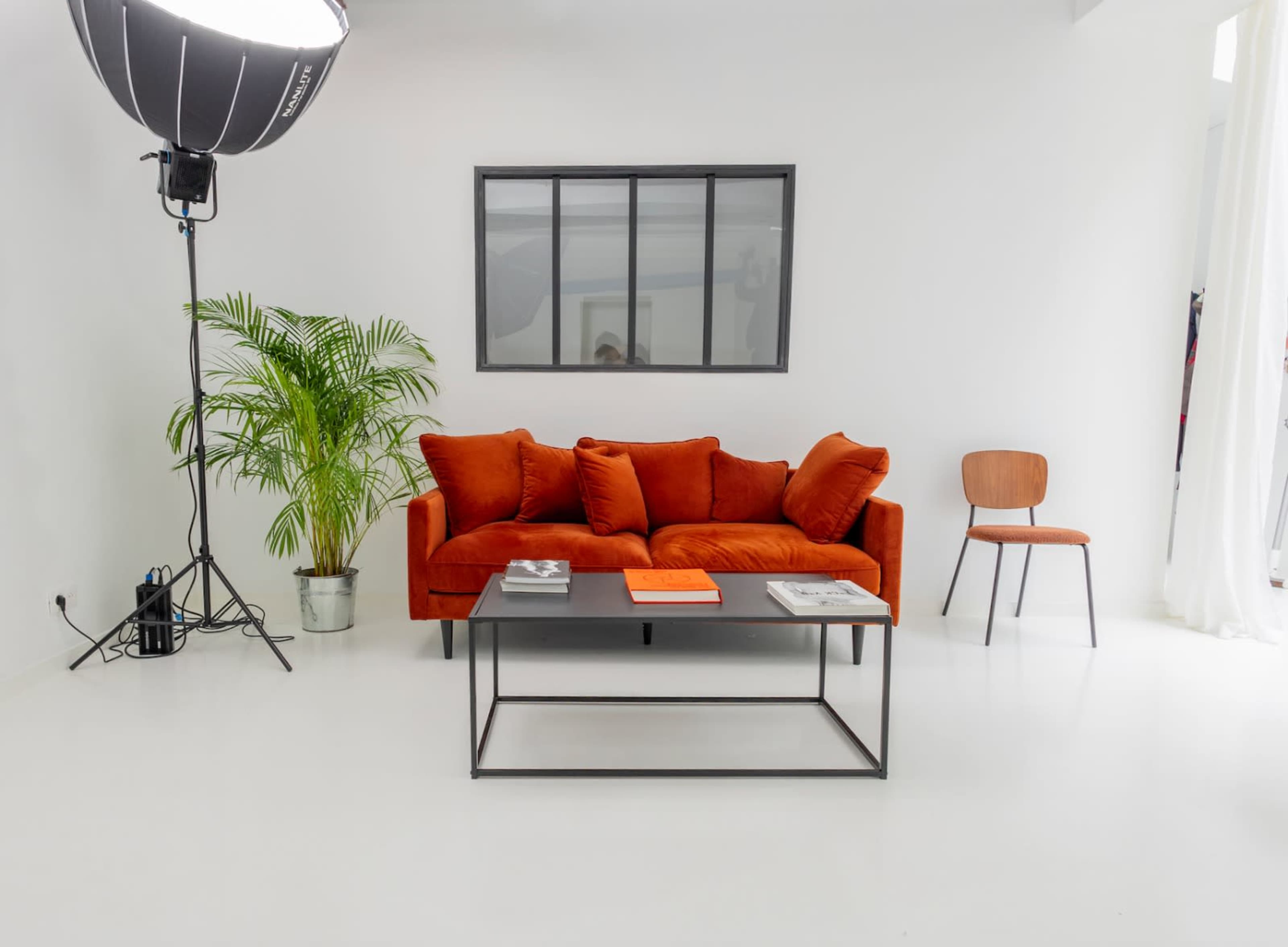 The image shows a modern living space featuring an orange sofa, a black coffee table with magazines, a potted plant, and a wooden chair, all against a white backdrop.