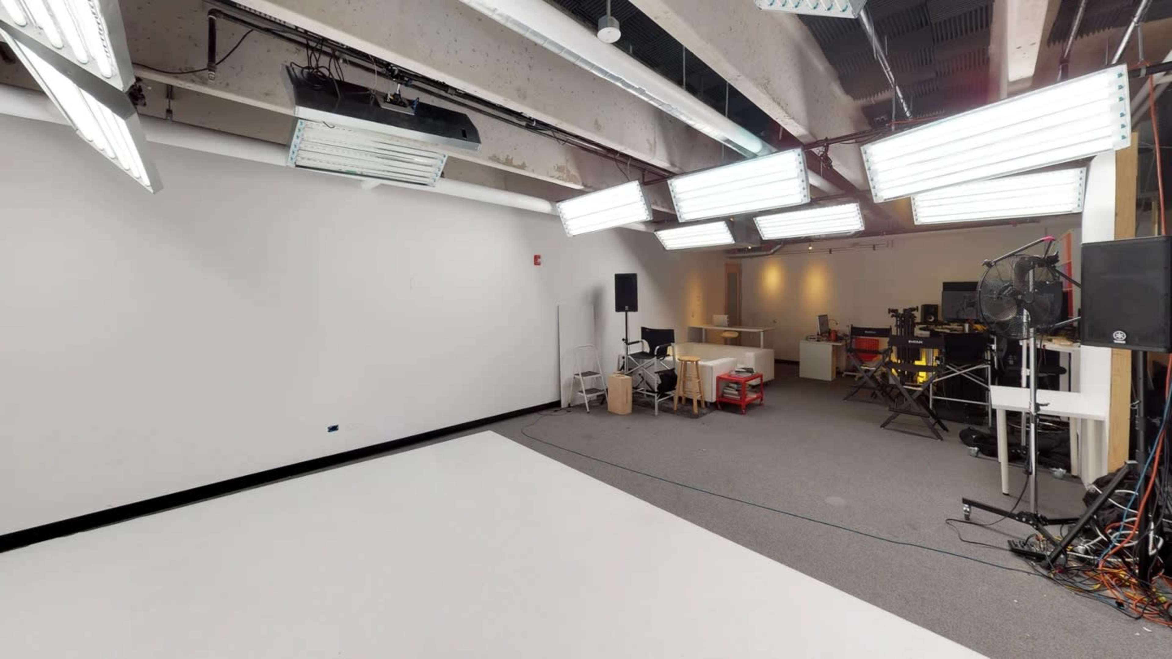 The image shows a brightly lit studio space with various lighting fixtures, a plain white backdrop, and equipment arranged along the walls.