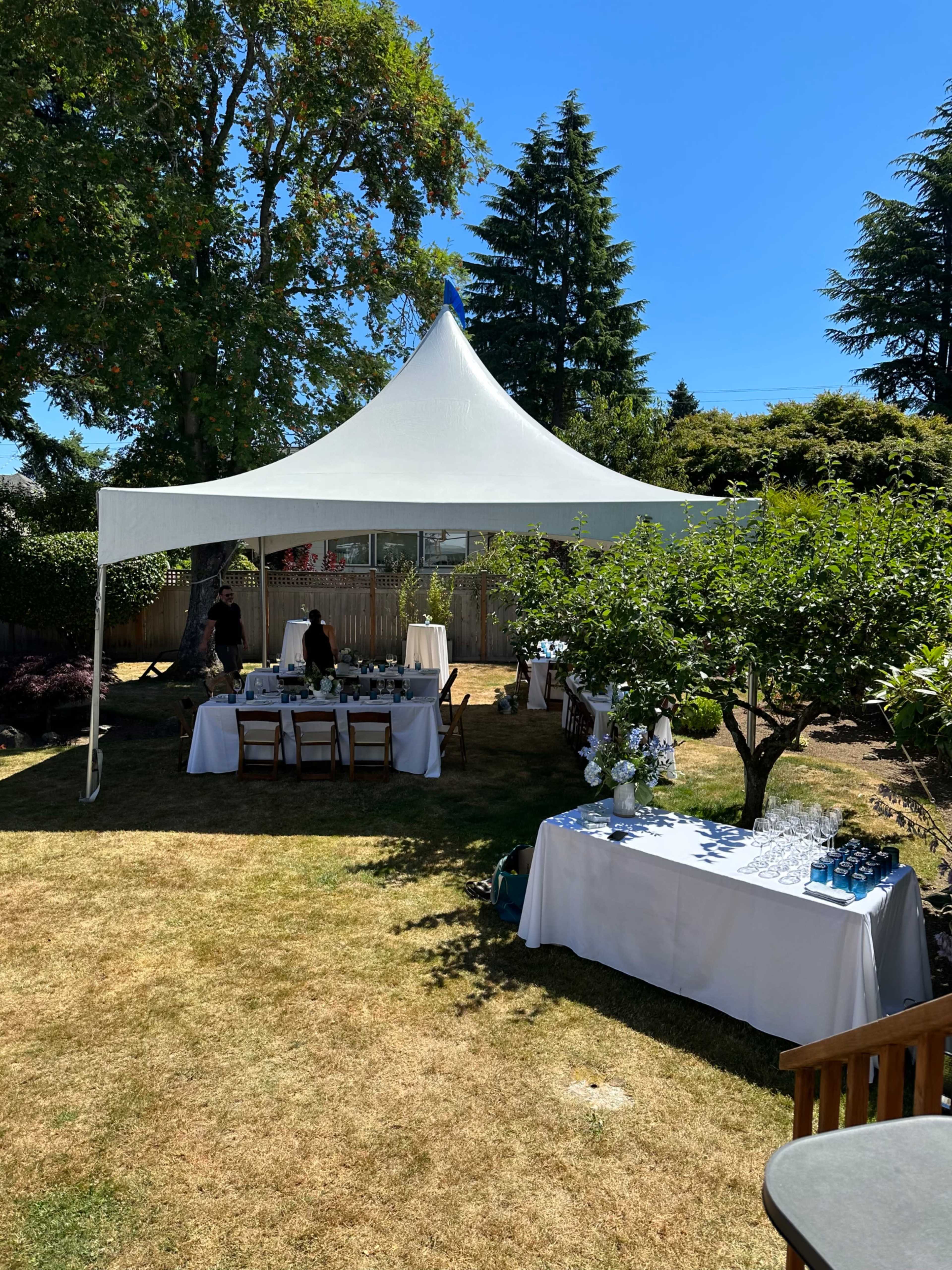 A white tent is set up in a grassy backyard, surrounded by trees, with tables arranged underneath it for an event.