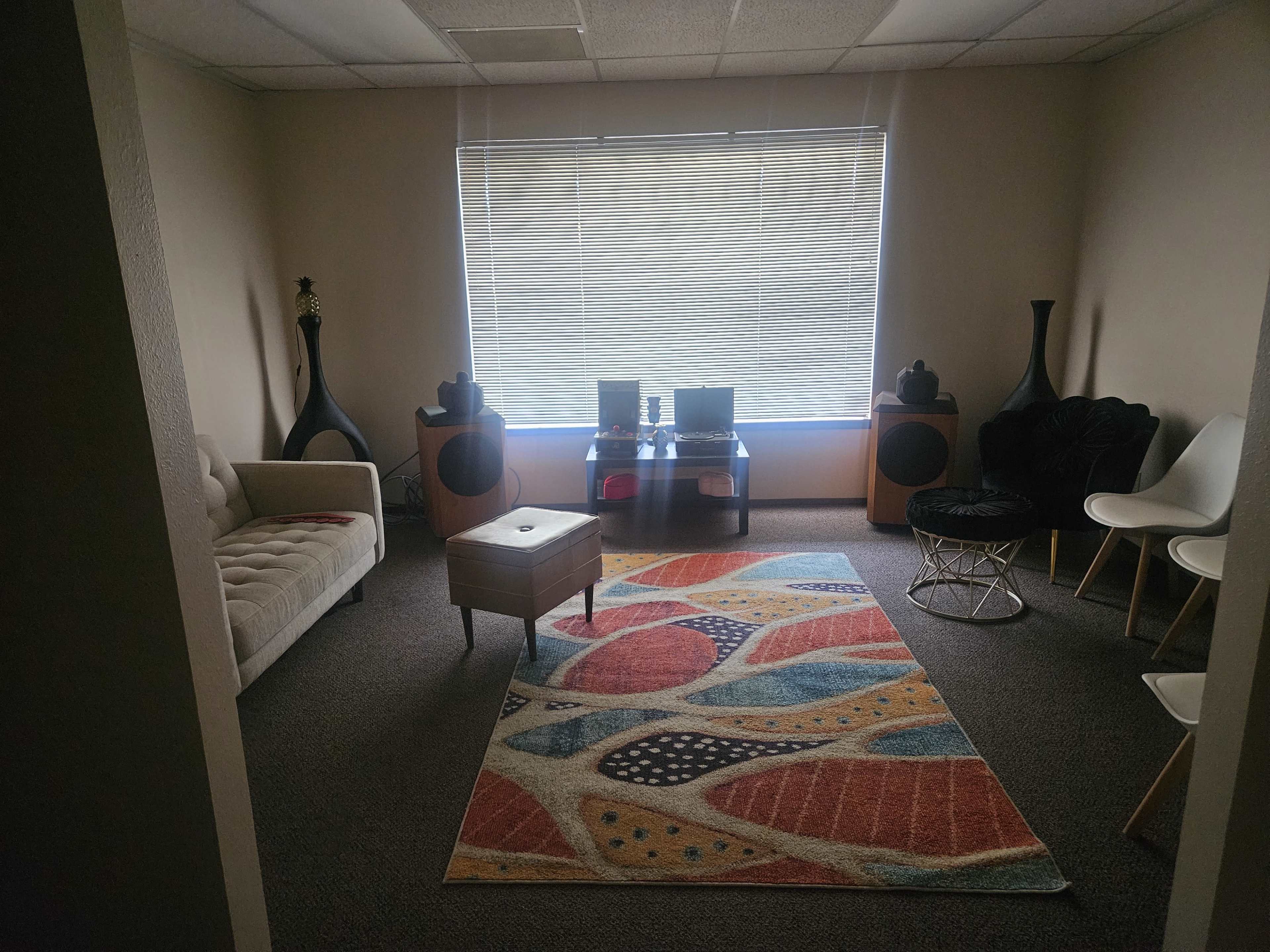The image shows a well-furnished office space with a couch, a coffee table, and several chairs arranged around a colorful rug.