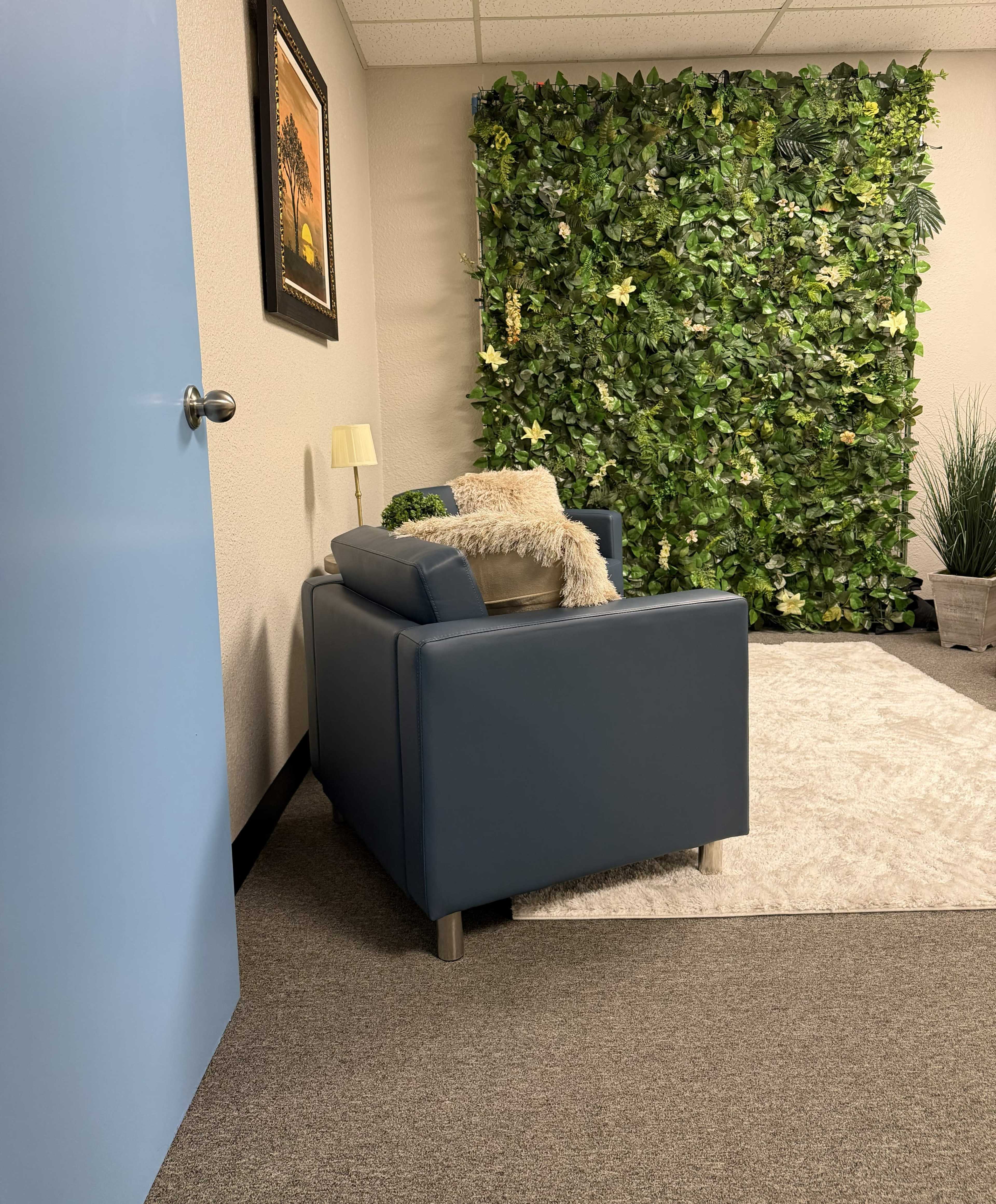 The image shows a small, well-decorated room featuring a blue armchair, a lamp, and a green plant wall.