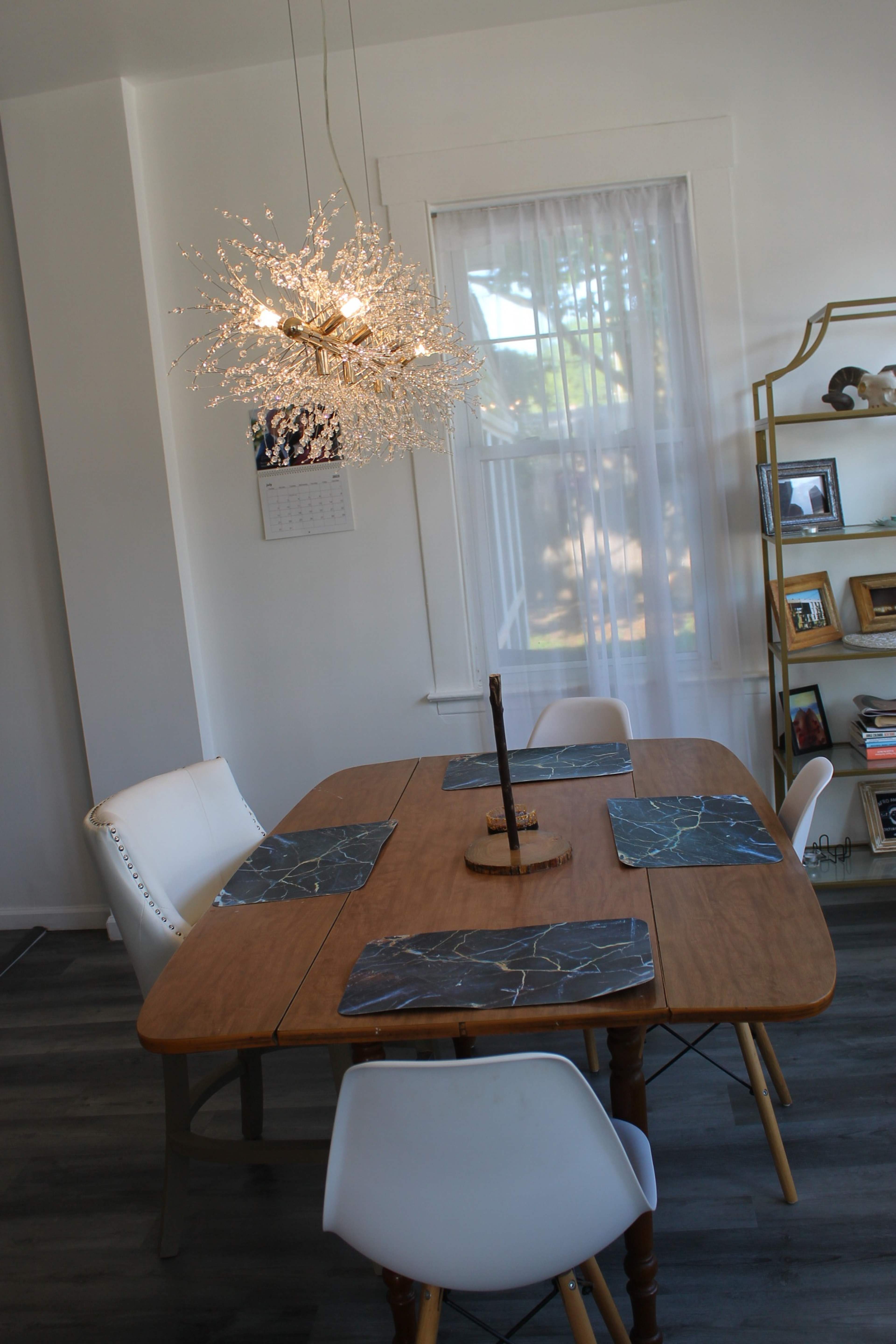 A wooden dining table with four chairs is set with placemats underneath a decorative light fixture and a nearby shelf holds framed photos.