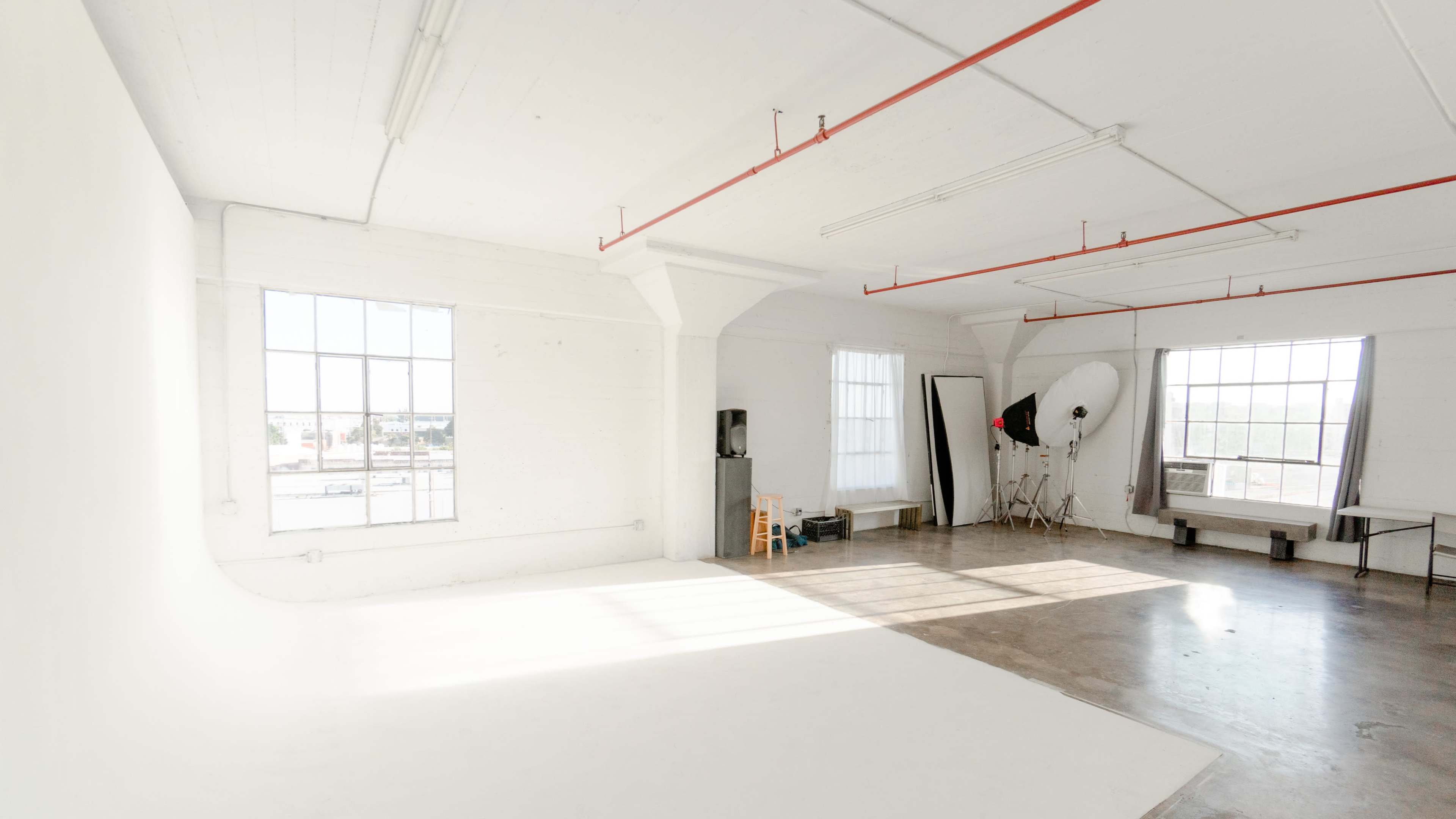 A spacious, well-lit photography studio featuring white walls, large windows, and various studio equipment.