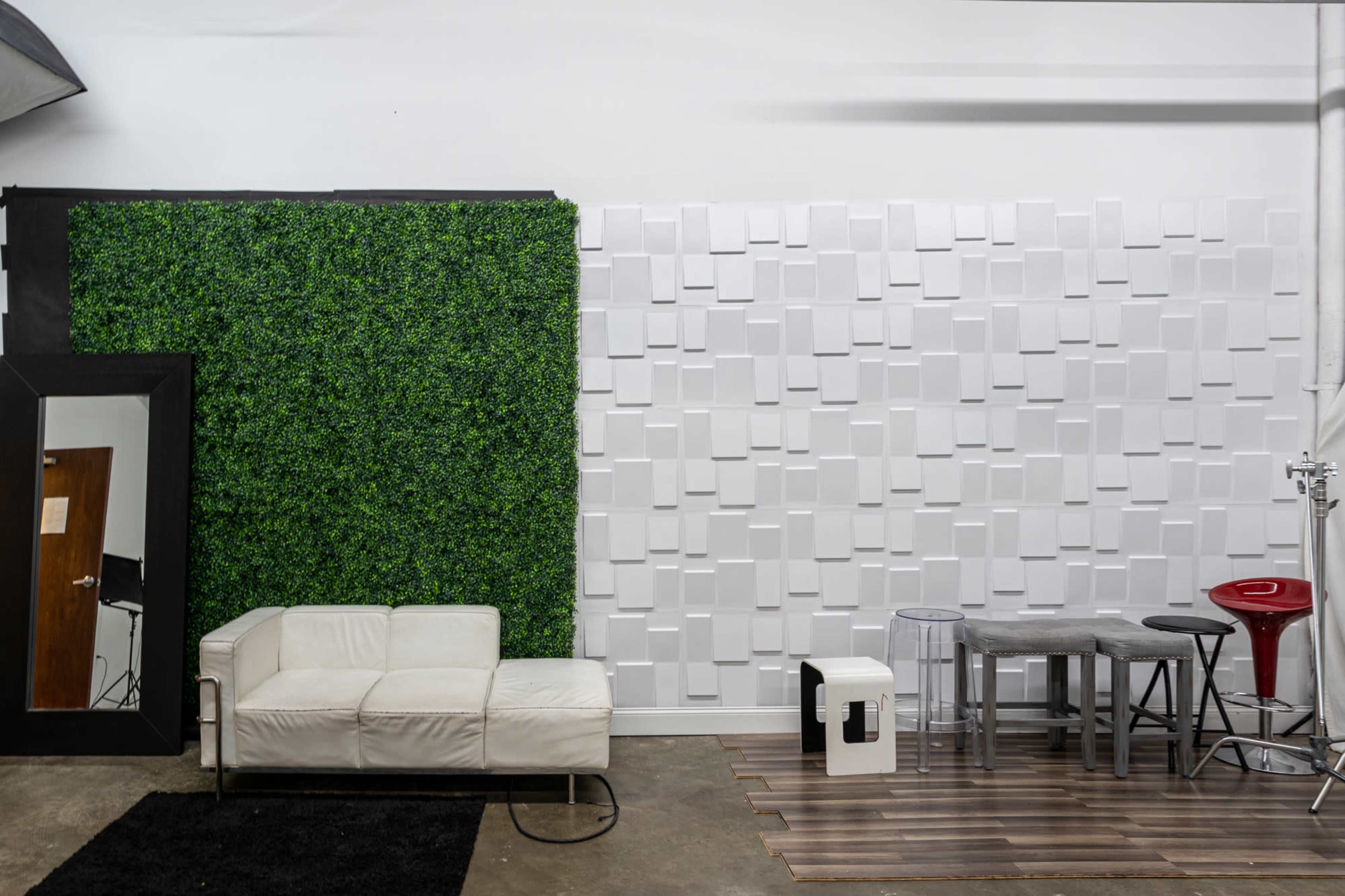 The image shows a room featuring a green wall of foliage, a white textured wall, a white couch, a wooden floor, and various pieces of furniture including a bar stool and a small table.