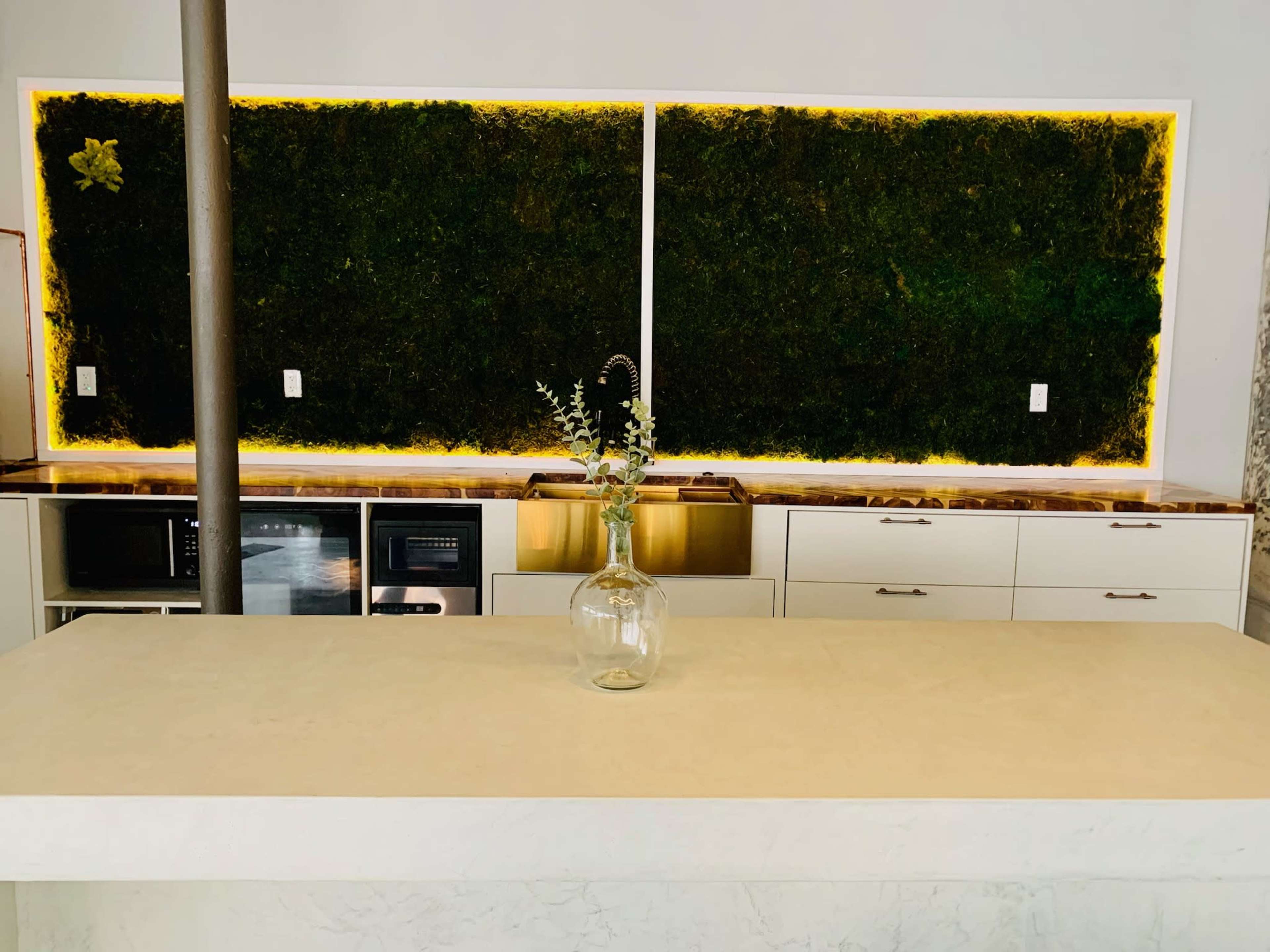 A modern kitchen with a large white island, cabinets below, and a vertical green moss wall illuminated by yellow lighting.