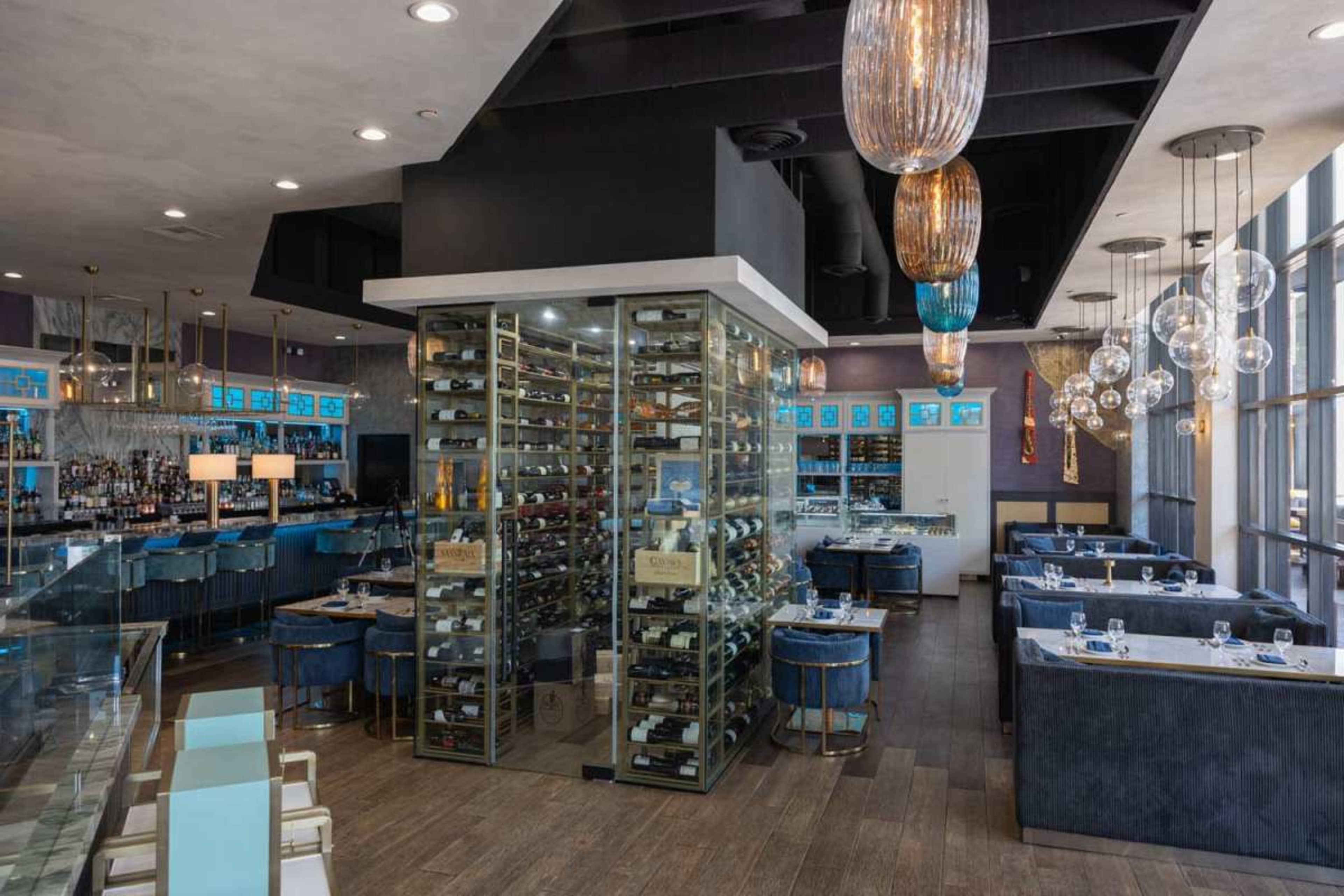 The image shows a modern restaurant interior featuring a wine storage area, blue upholstered seating, and pendant lighting.