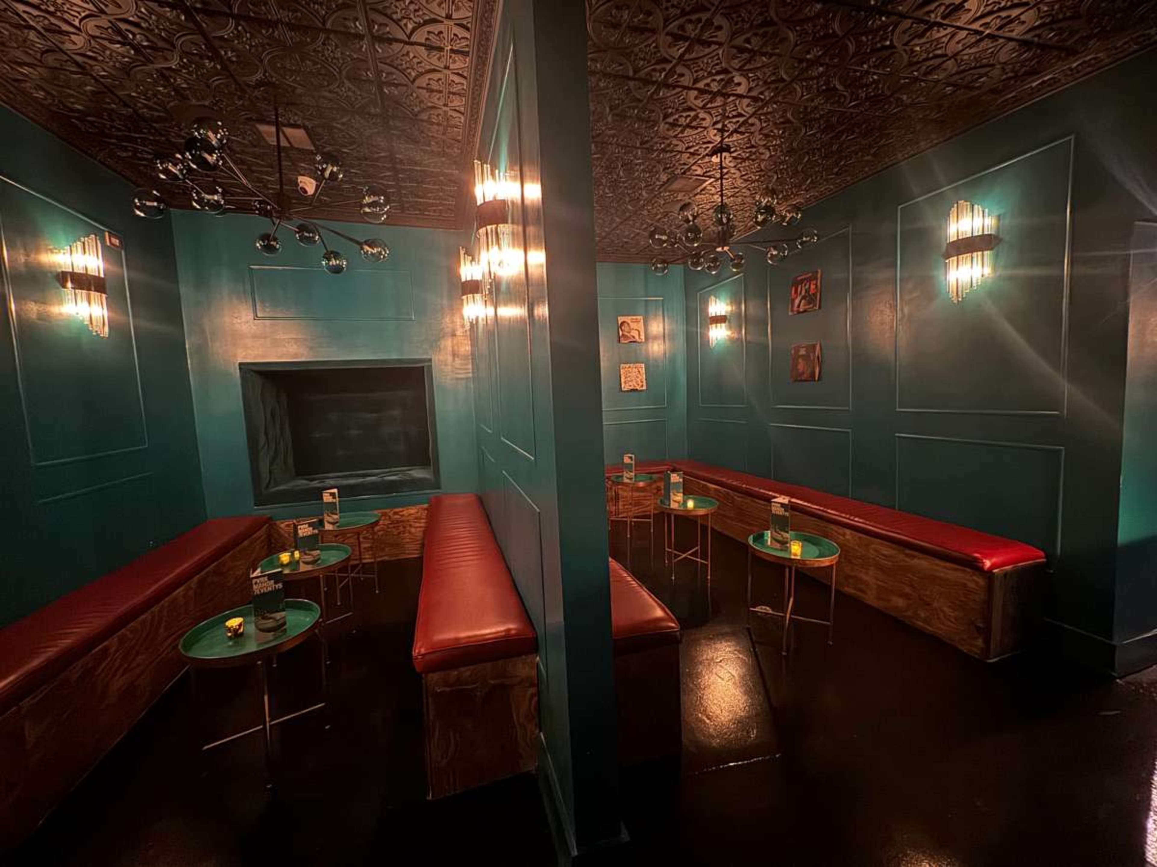 The image shows a dimly lit lounge with teal walls, red seating, and small round tables, featuring a central dividing wall and decorative lighting.