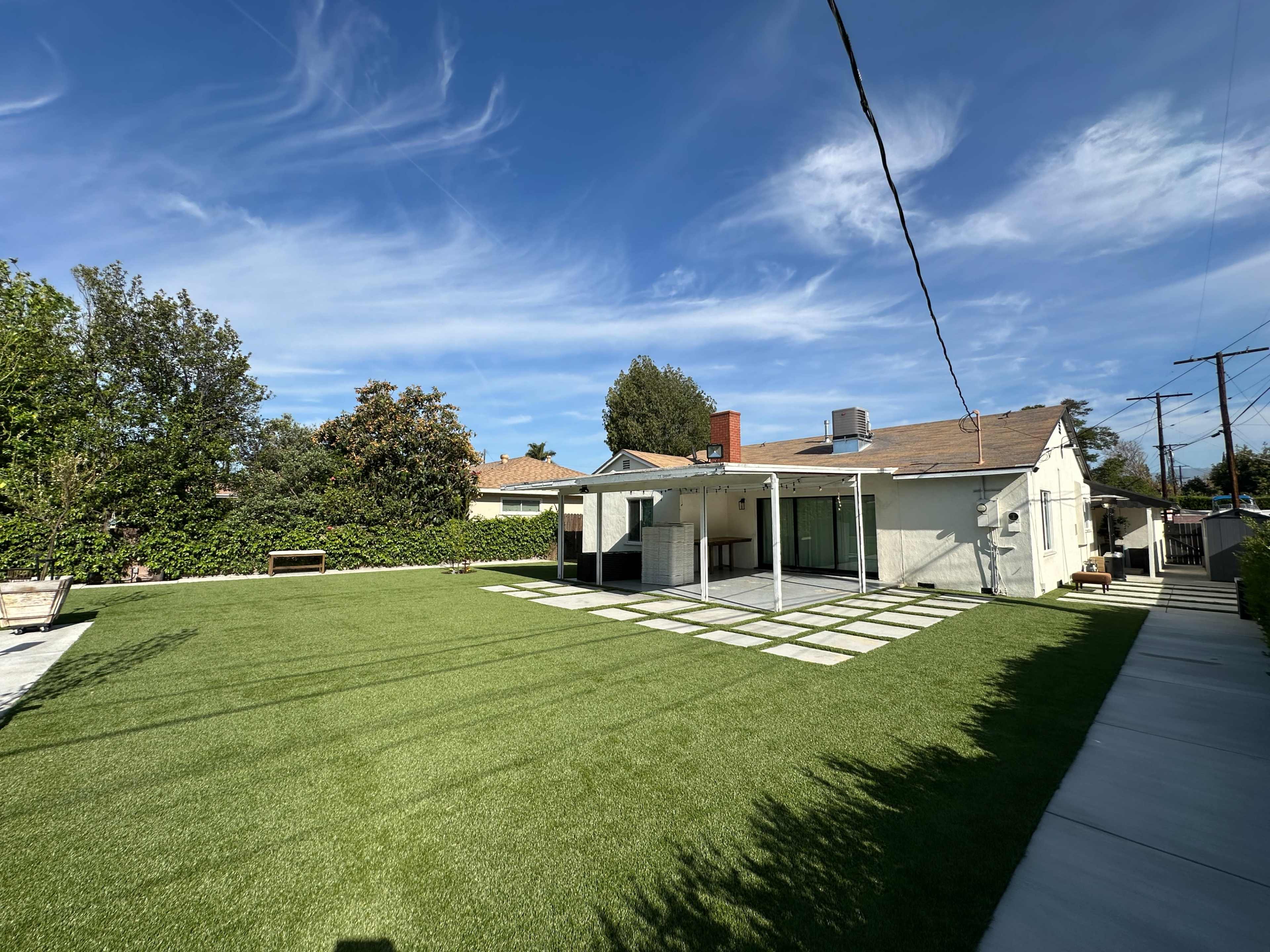 The image shows a sunny backyard with artificial grass, a patio area, and a single-story house in the background.
