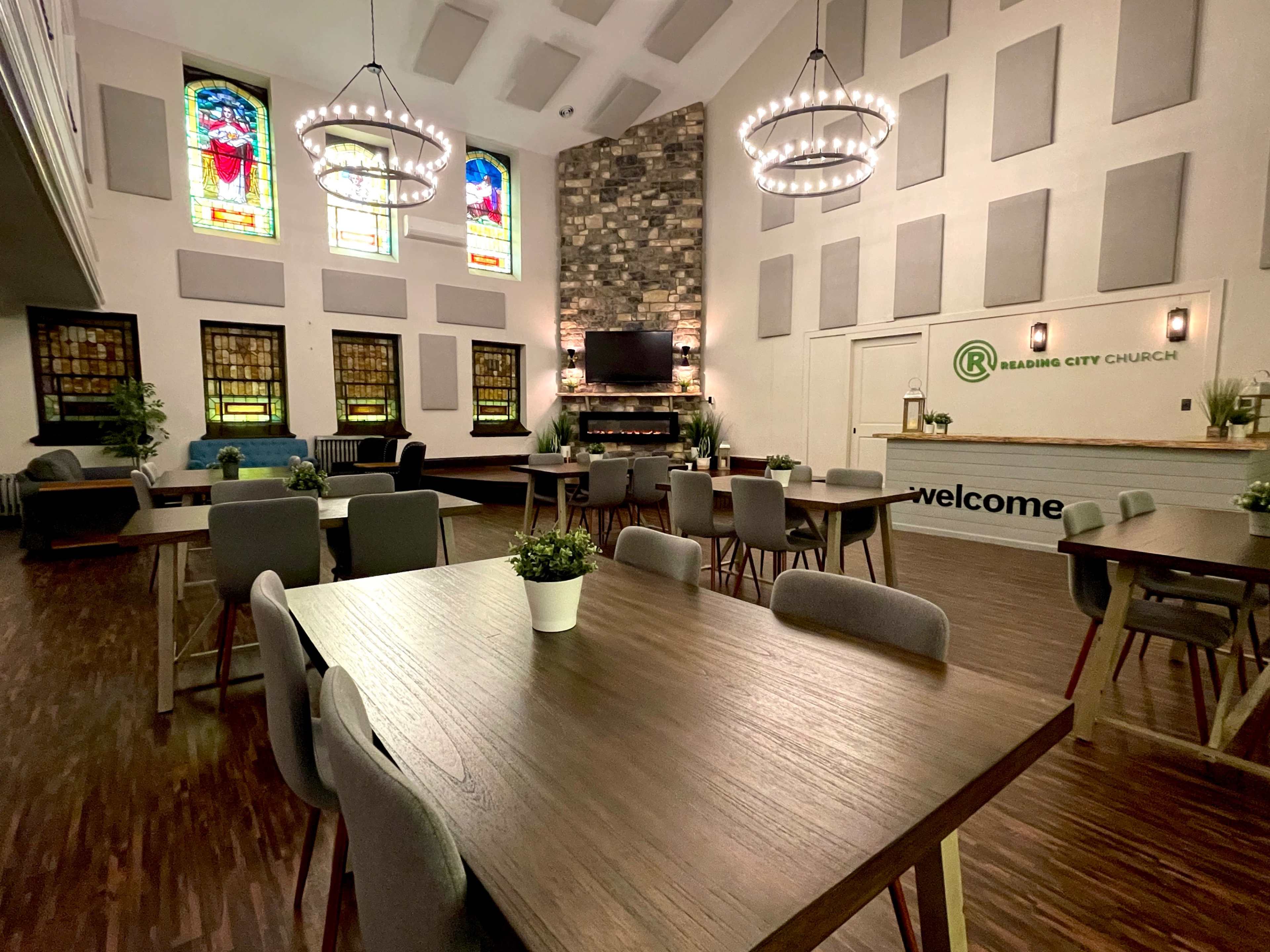 The interior of a church features tables and chairs set up for gatherings, with stained glass windows and a stone fireplace as focal points.