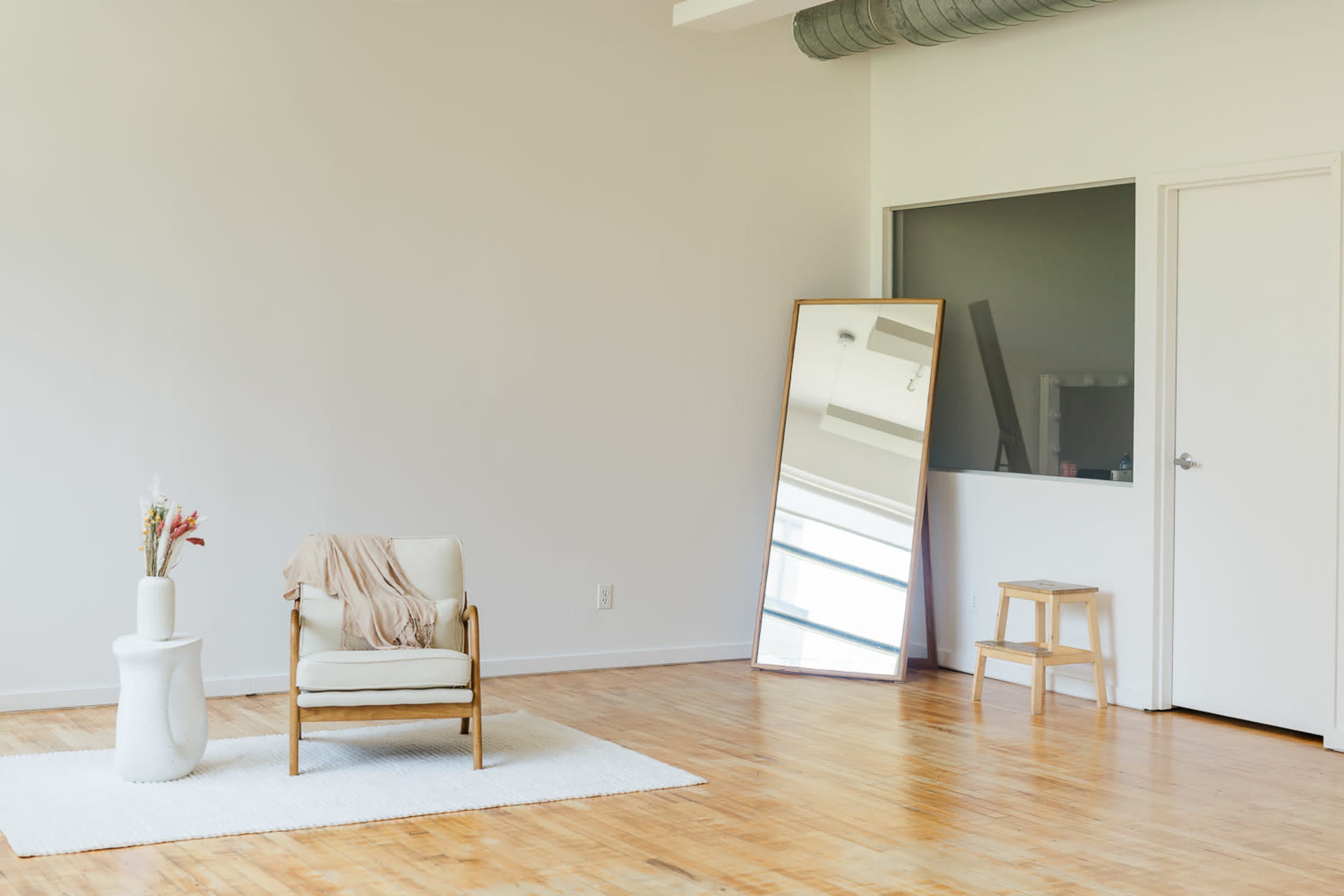 The image features a minimalistic interior space with a light wooden floor, a white rug, a single armchair, a small table with flowers, a tall mirror, and a stool.