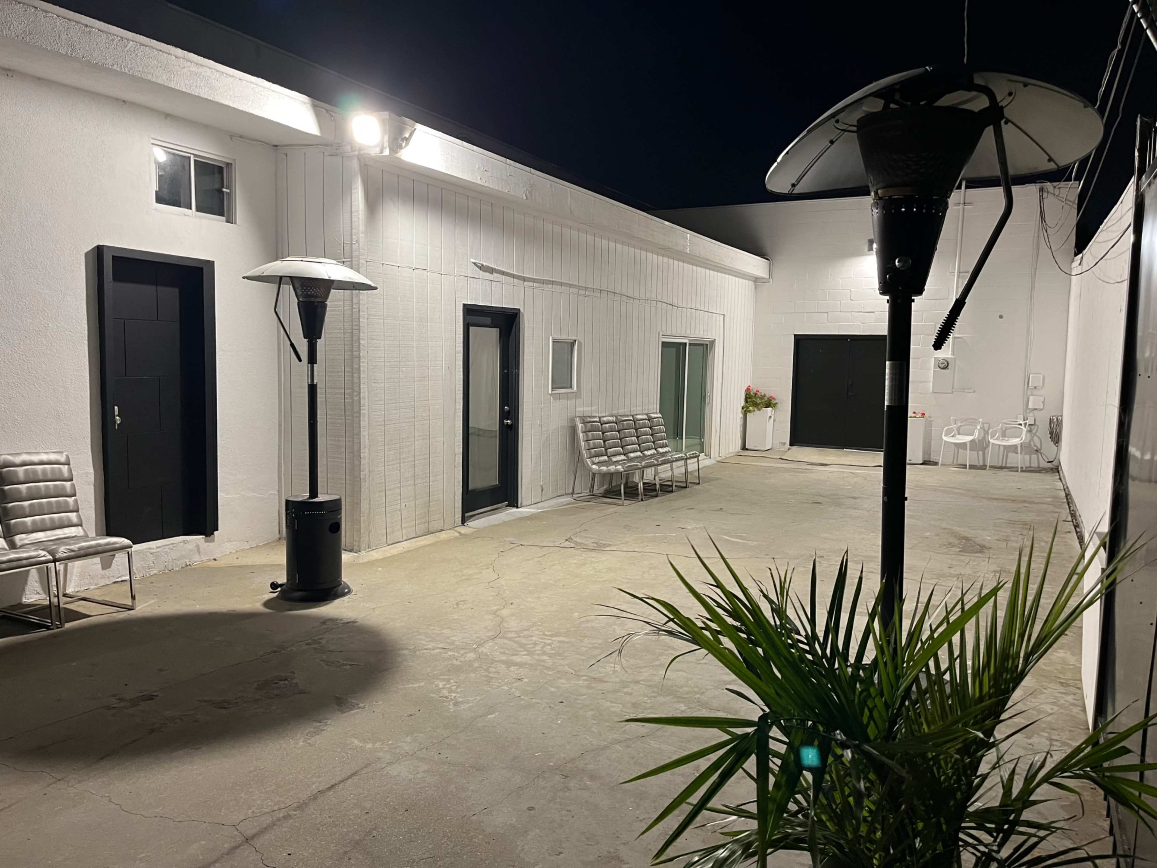 The image shows a dimly lit outdoor area with several metal chairs, a plant in the foreground, and light fixtures illuminating the white walls of the surrounding buildings at night.
