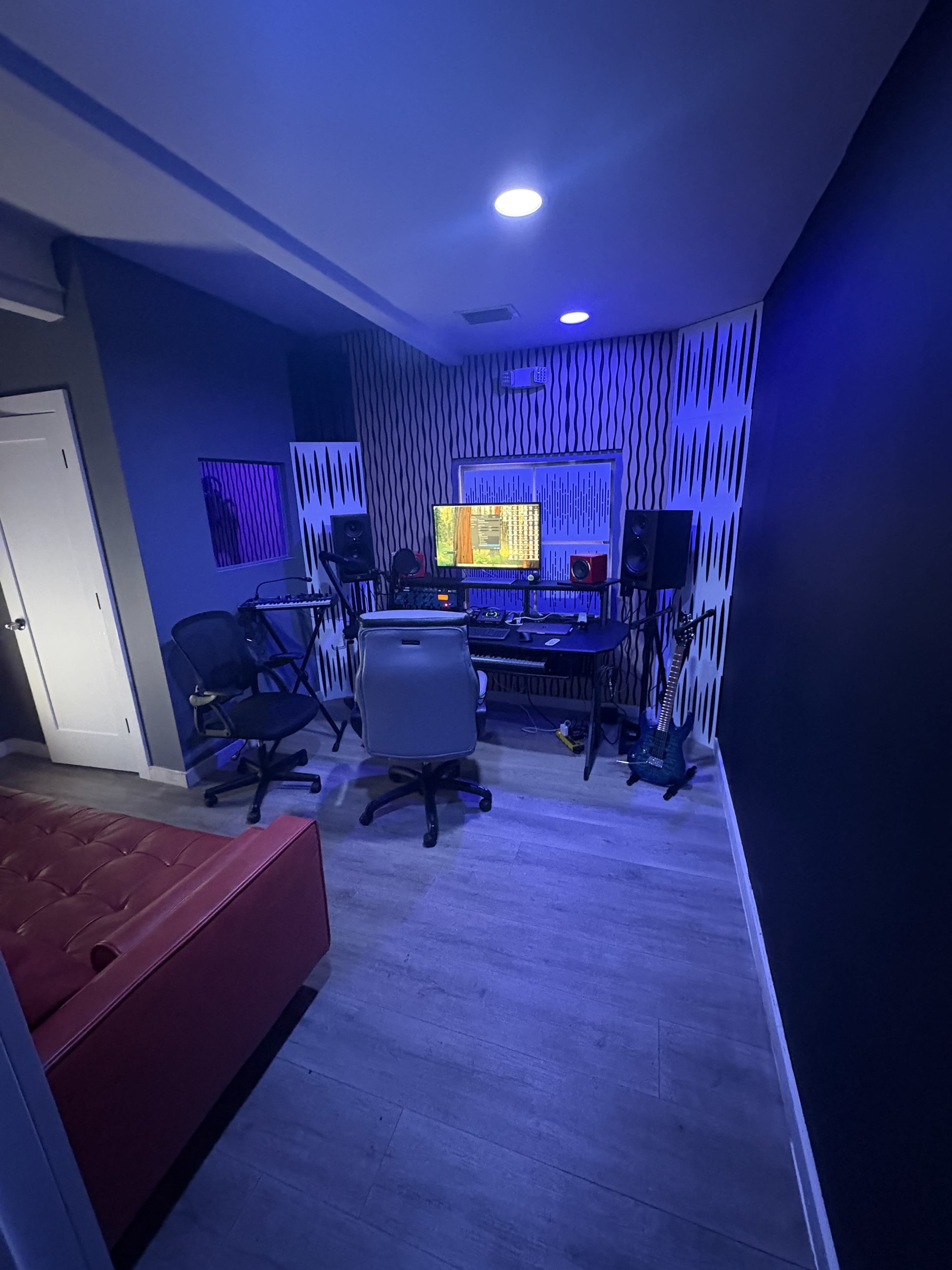 A modern recording studio features music production equipment, a computer monitor displaying audio software, and chairs arranged around a desk, with purple ambient lighting.