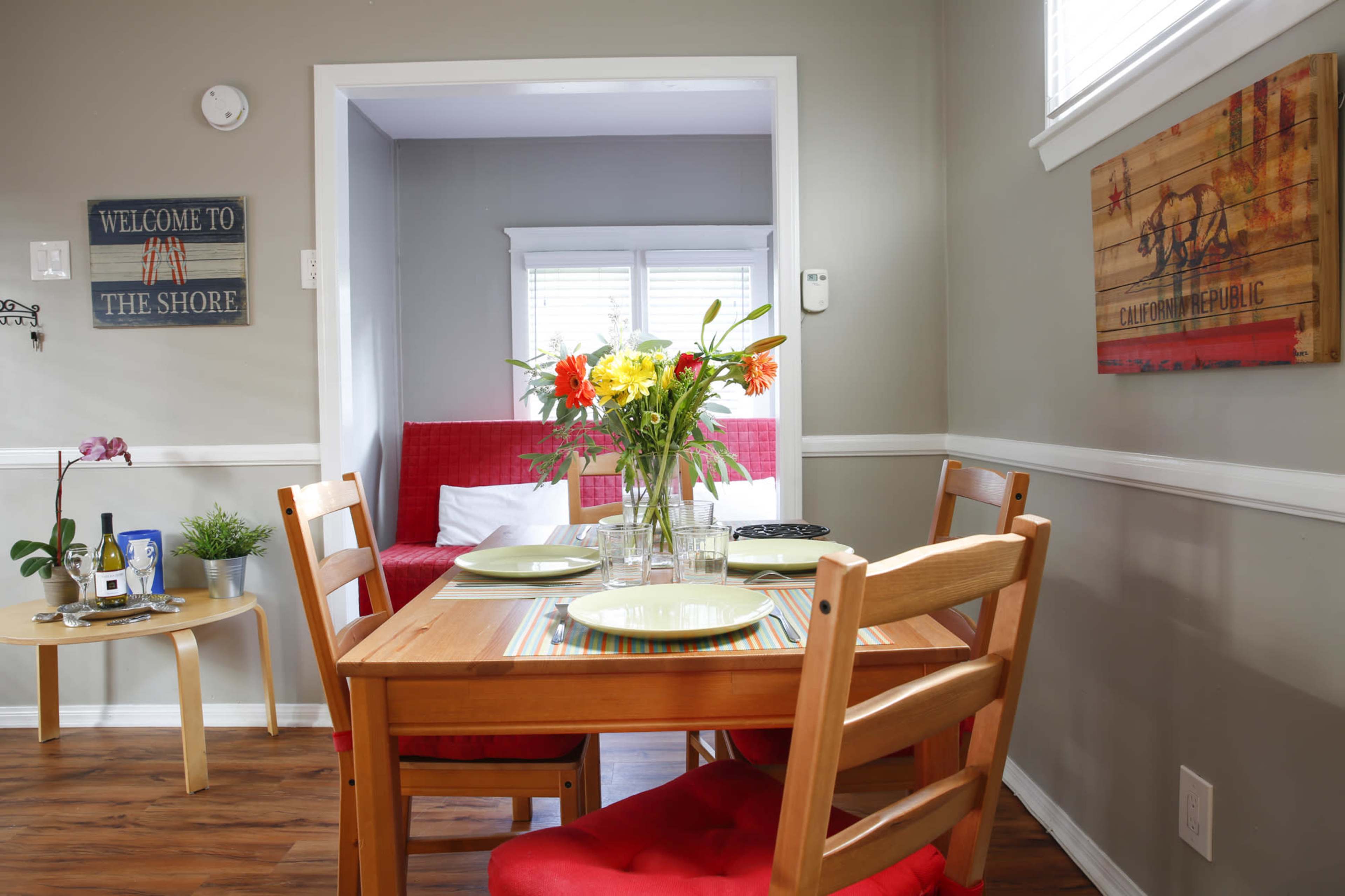 A small dining area features a wooden table set for a meal, surrounded by chairs, with a colorful vase of flowers as the centerpiece and a cozy living space visible in the background.