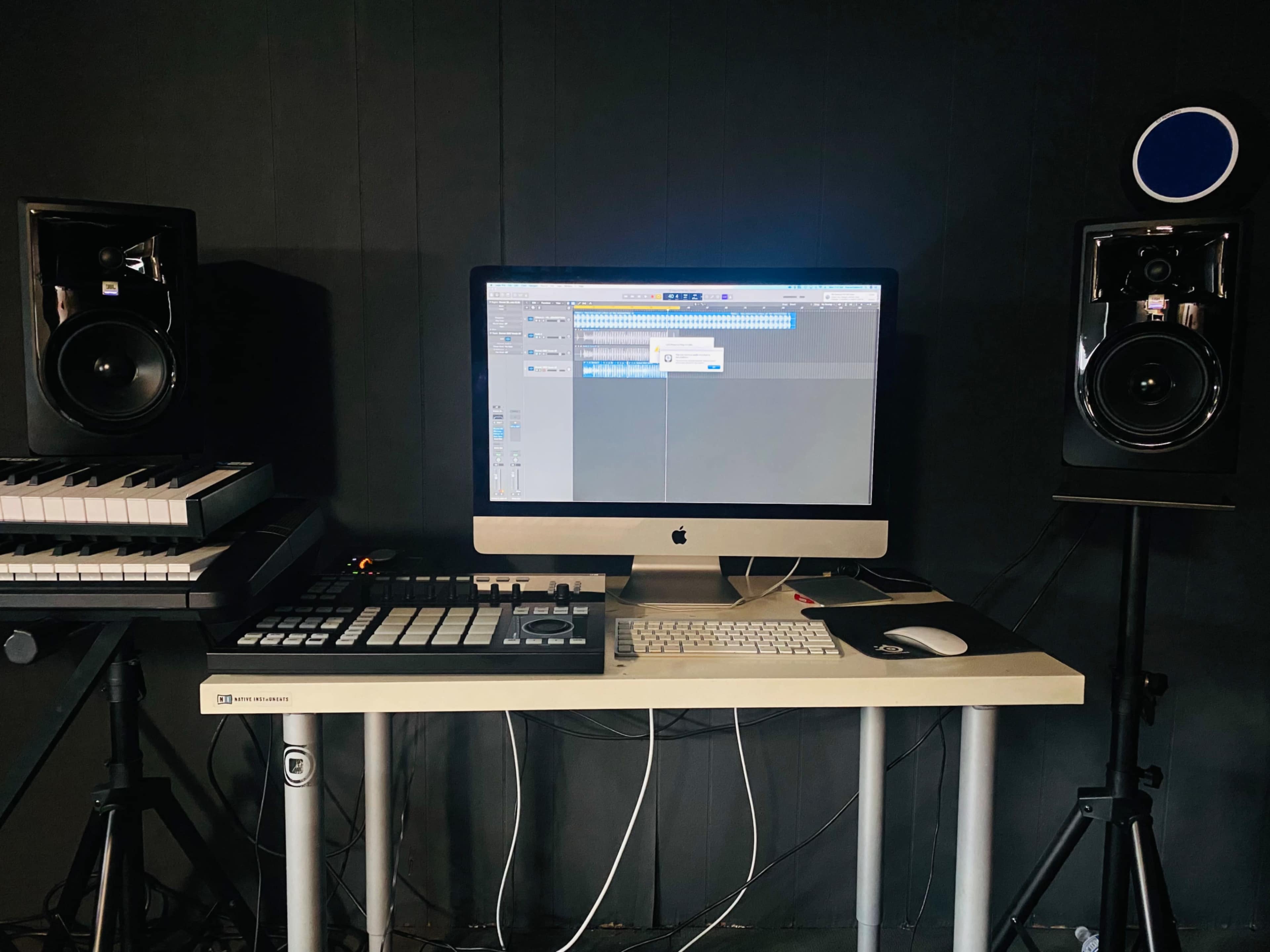 A music production workspace featuring an iMac with a digital audio workstation open, flanked by studio monitors and a MIDI controller on a desk.