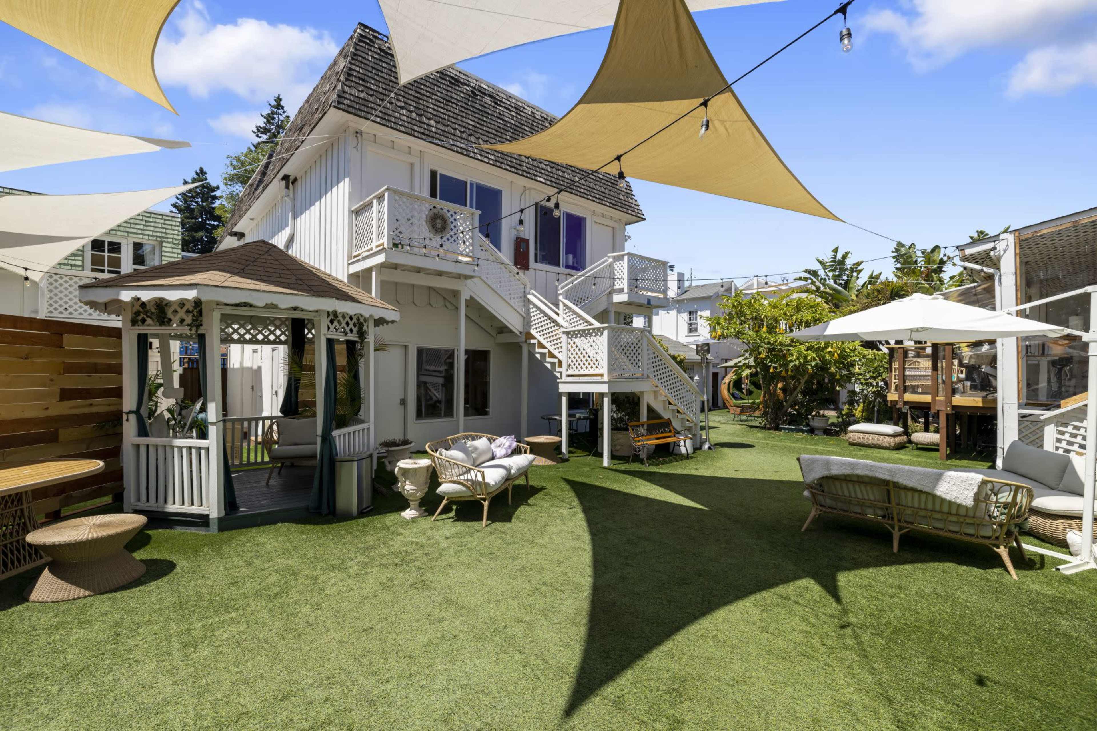 The image shows a spacious outdoor area featuring a two-story house, numerous seating arrangements, and decorated with fabric canopies overhead.