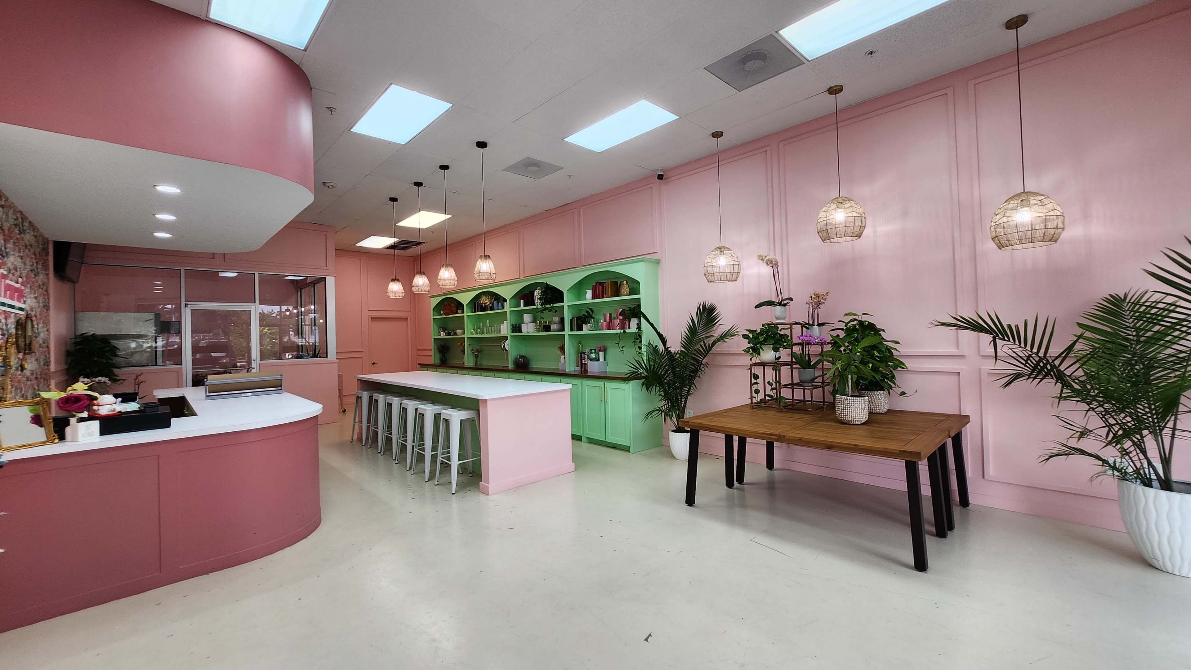 A brightly lit interior space featuring pink and green walls, a central island with white stools, and several plants and hanging lights.