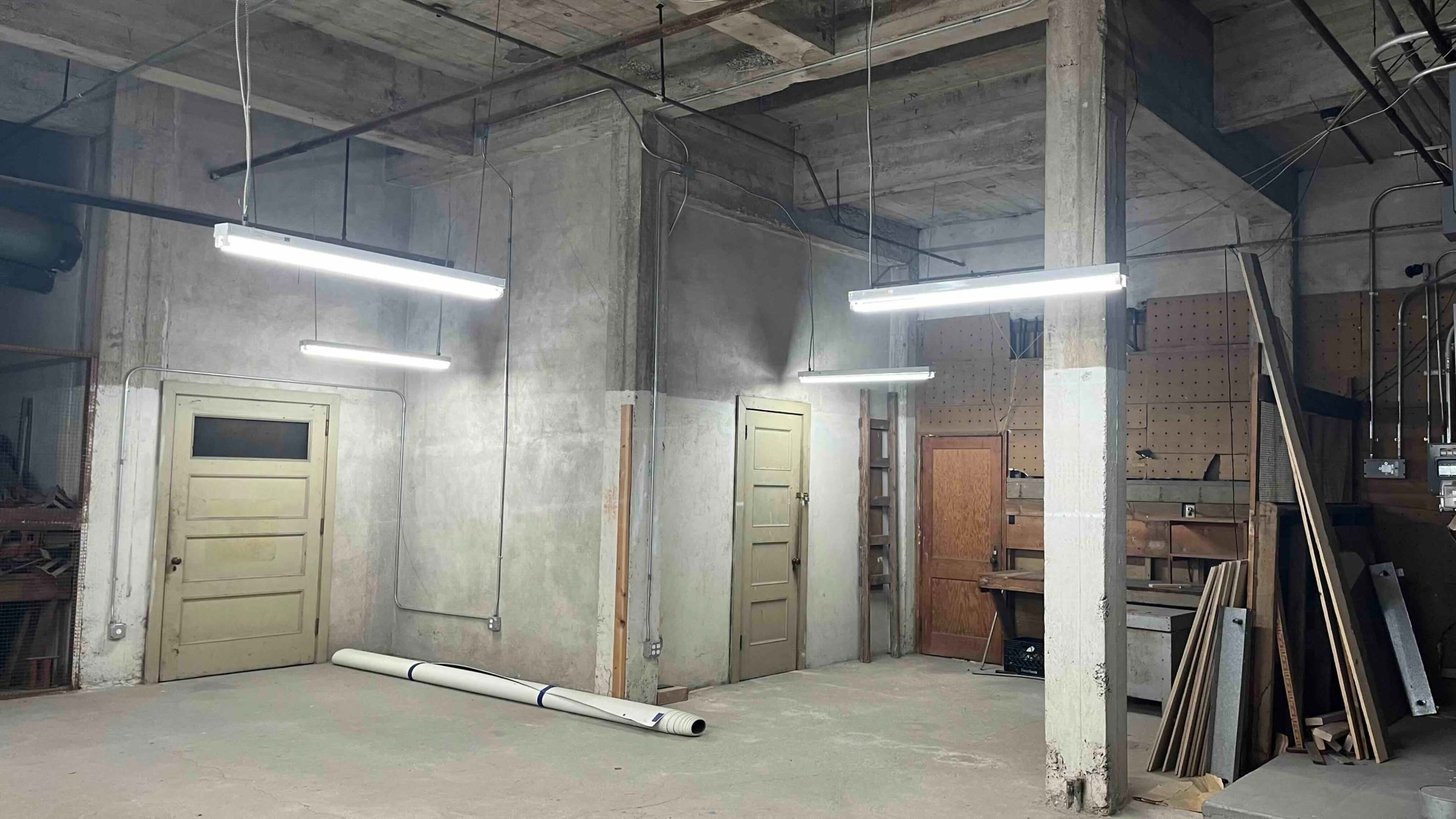 The image shows a dimly lit room with concrete walls, featuring two closed doors on one side, a workbench with tools on another, and a rolled-up white pipe on the floor.
