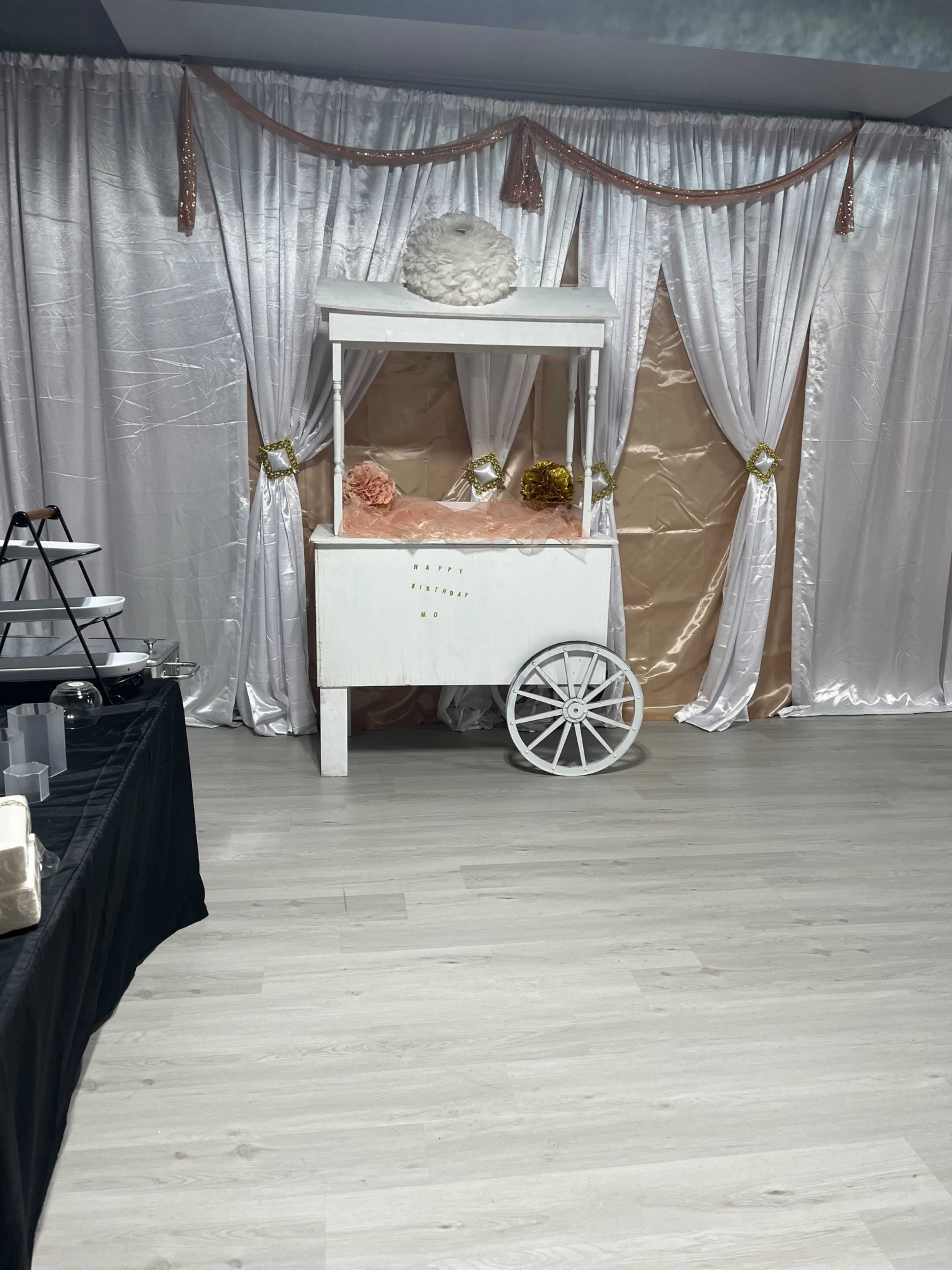 A decorative cart with a white wooden structure and pink decorative elements, set against a backdrop of draped satin fabric and an elegant floor.