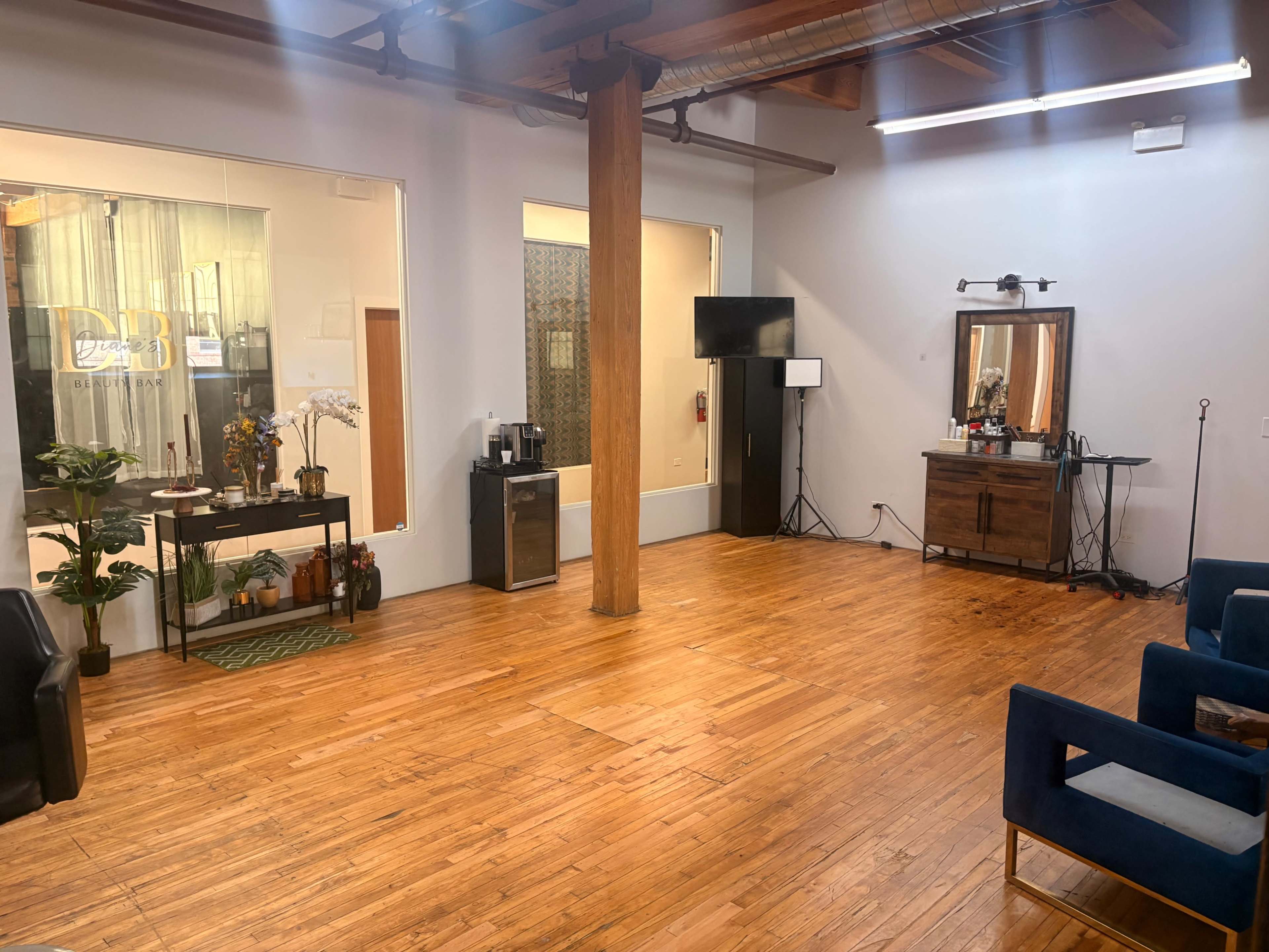The image shows an open, well-lit room with wooden flooring, featuring a seating area with blue chairs, a television, and a decorative table against one wall.
