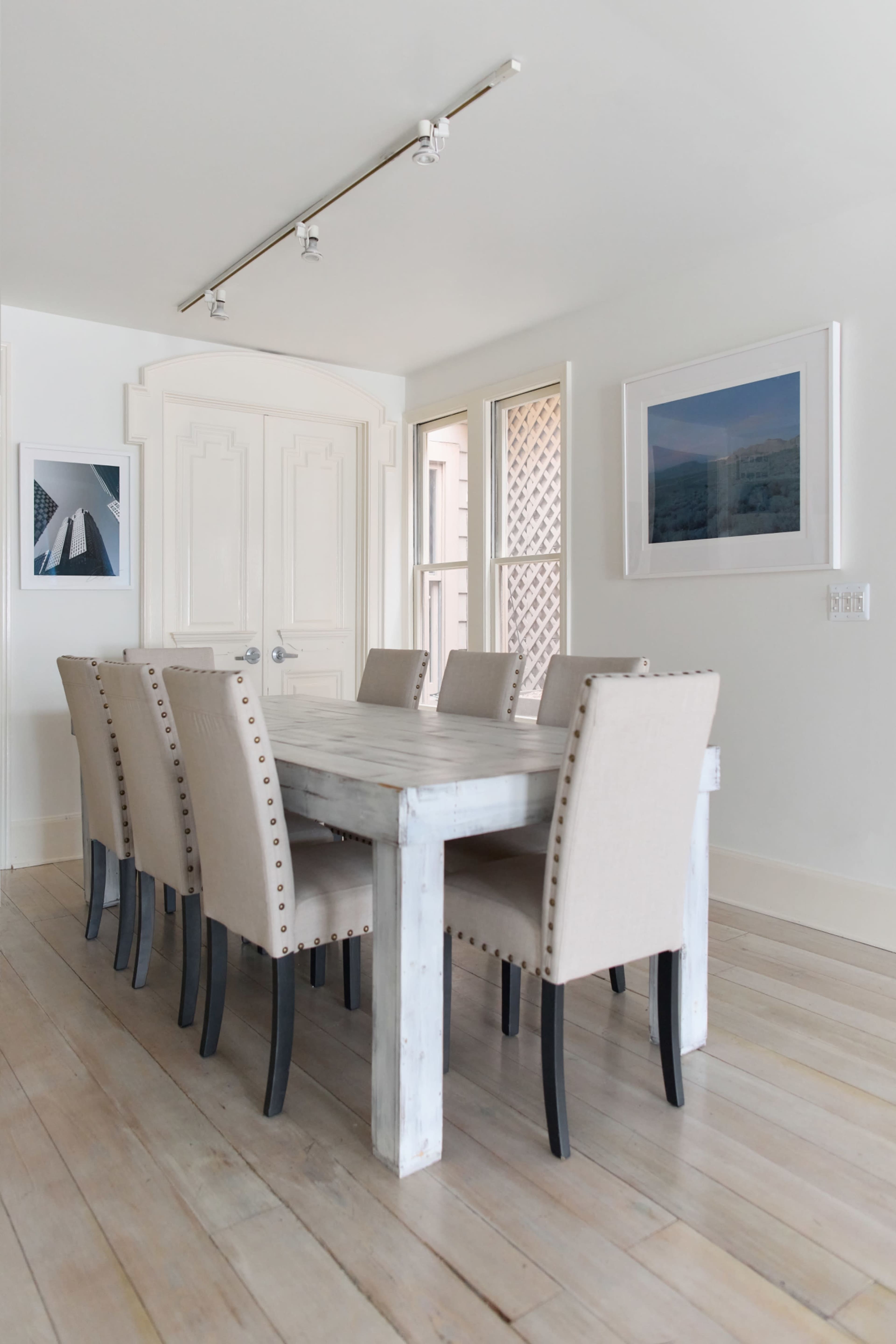 A long wooden dining table is surrounded by beige upholstered chairs in a bright, minimalist room with white walls and art on the walls.