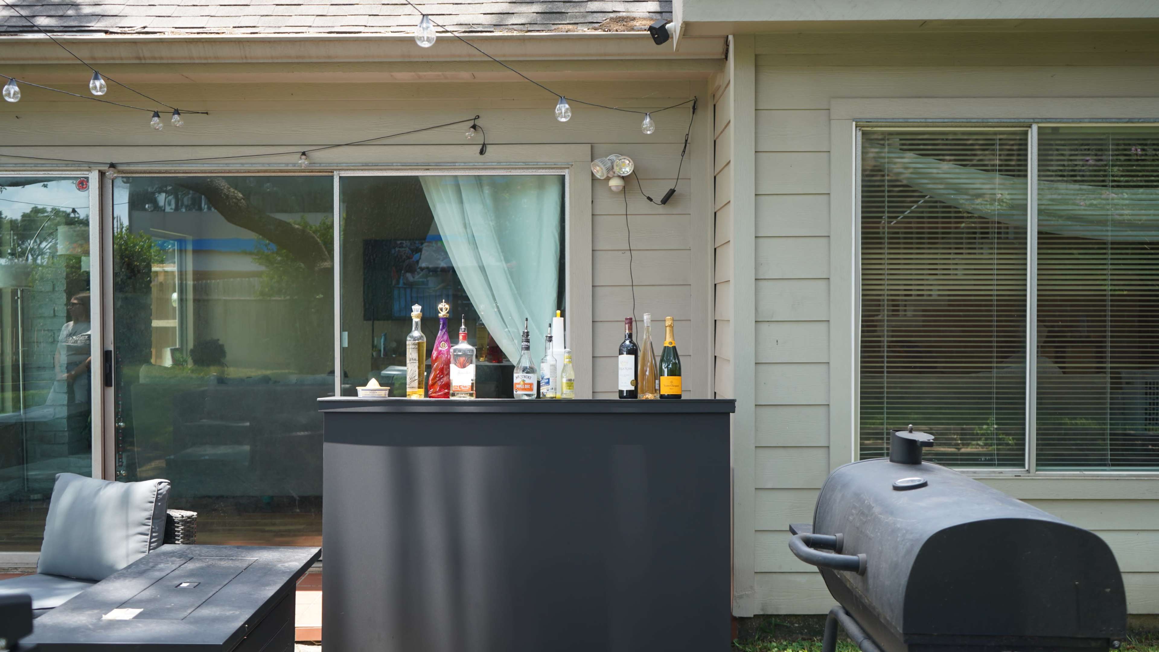 The image shows a backyard setup with a black bar featuring various bottles of liquor, along with a grill and a patio area in front of a house.