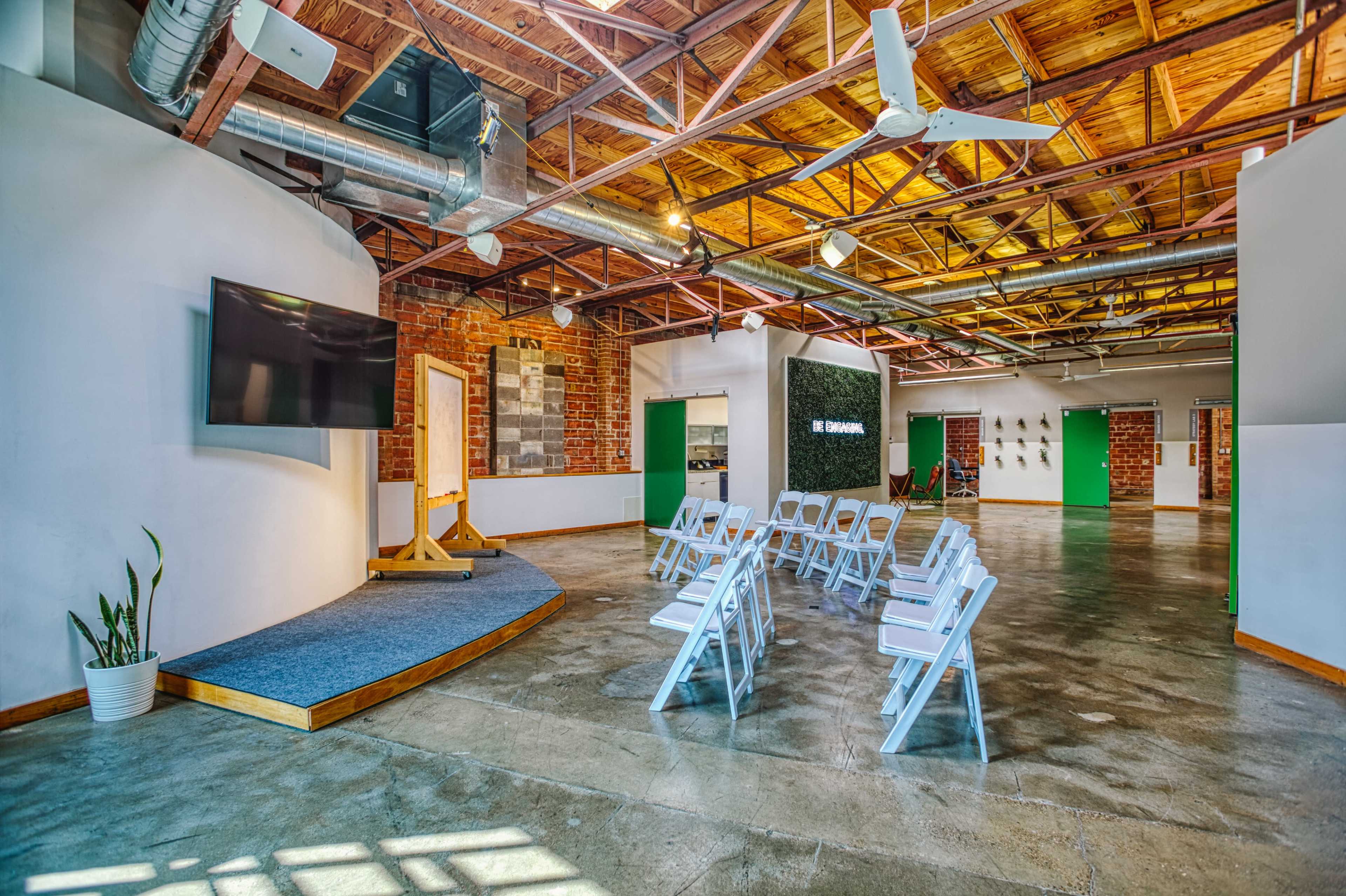 The image shows a spacious interior with wooden beams, featuring a presentation area with a screen and rows of white chairs arranged for an event.