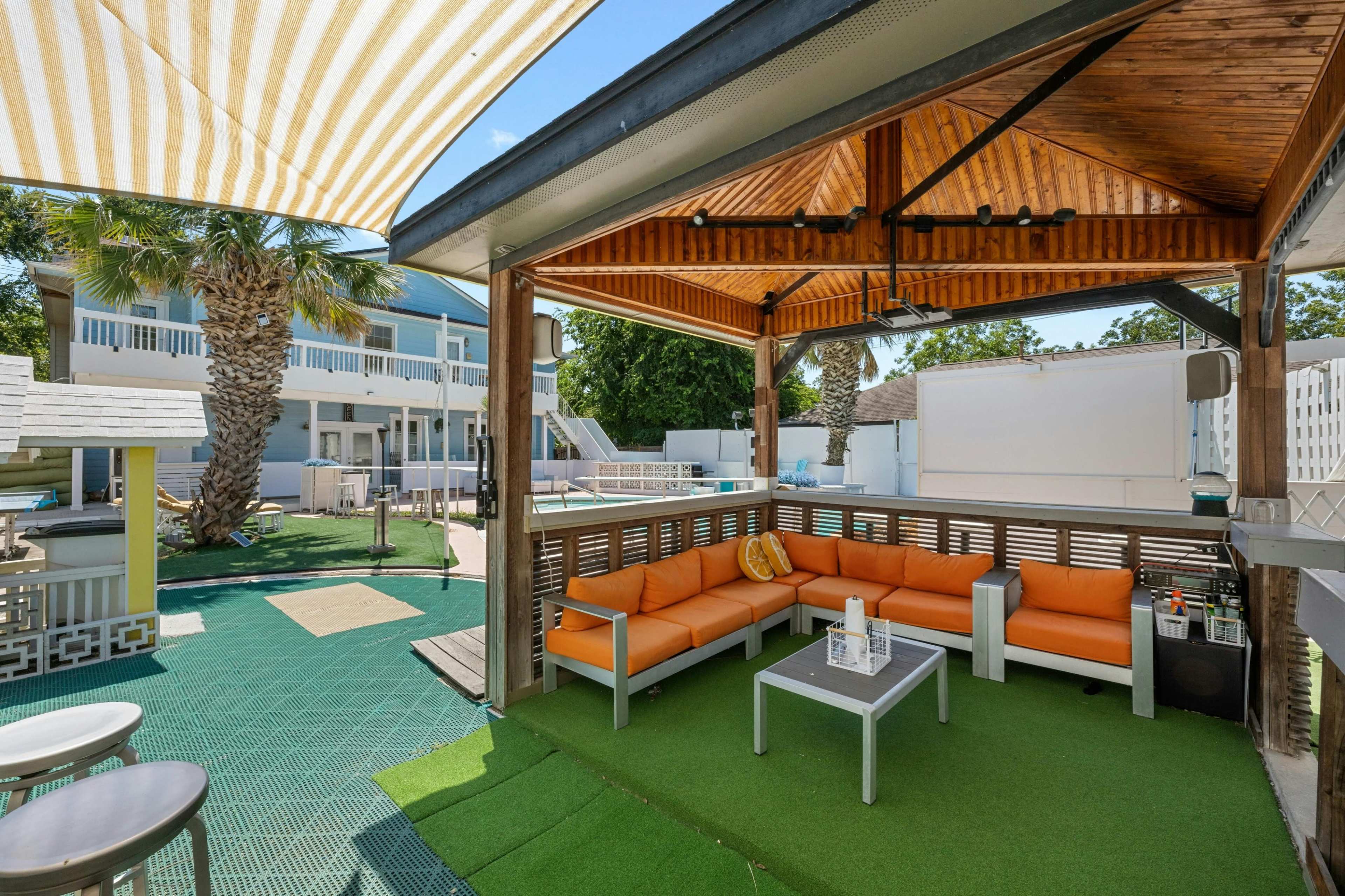 The image shows a covered outdoor seating area with orange couches and a small table, surrounded by green lawn and palm trees.