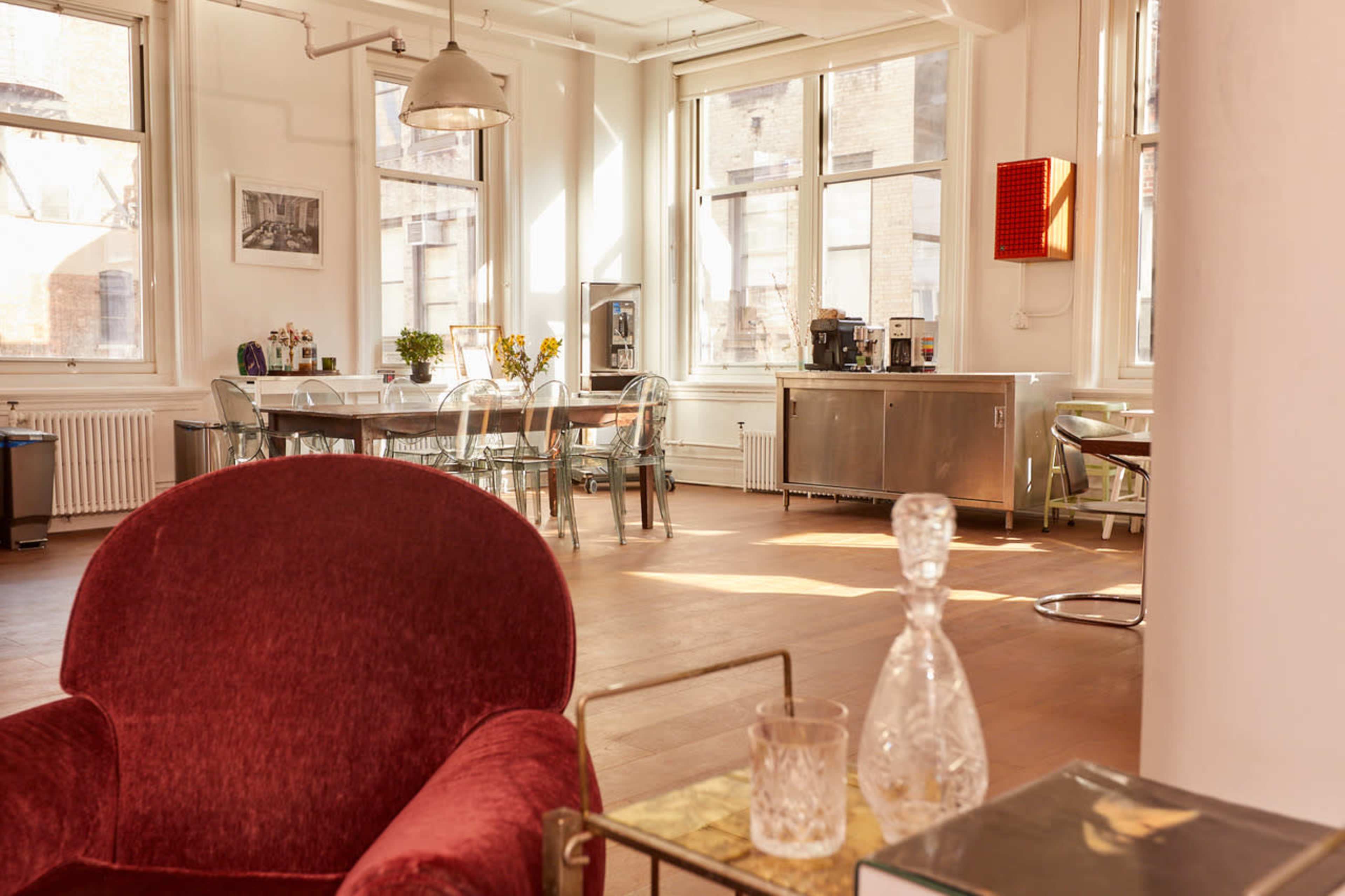 The image shows a spacious, sunlit room with a red armchair in the foreground, a dining table surrounded by clear chairs, and large windows allowing natural light to fill the space.