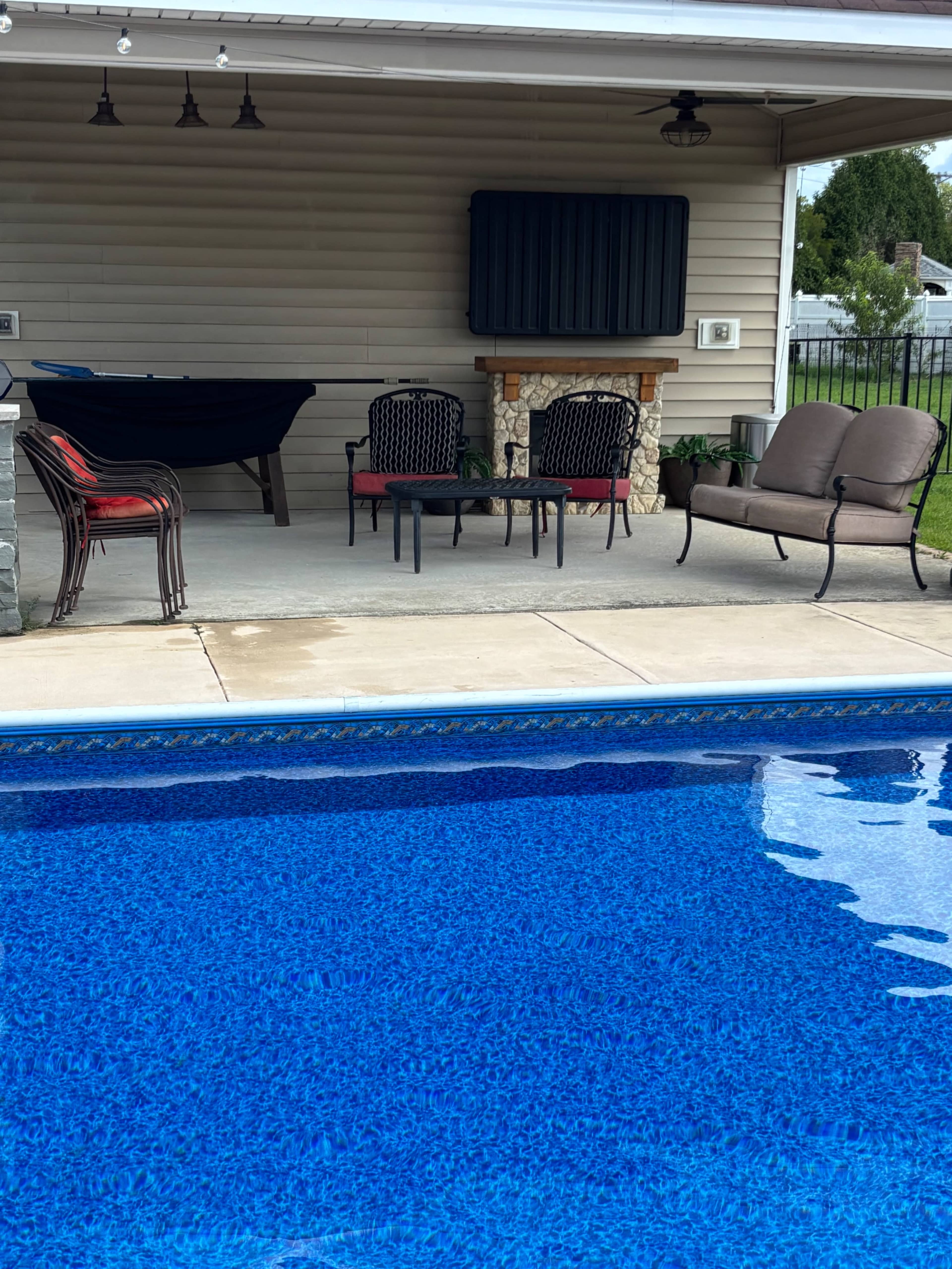 The image shows a swimming pool in the foreground with clear blue water, and a shaded patio area that includes a table and chairs in the background.