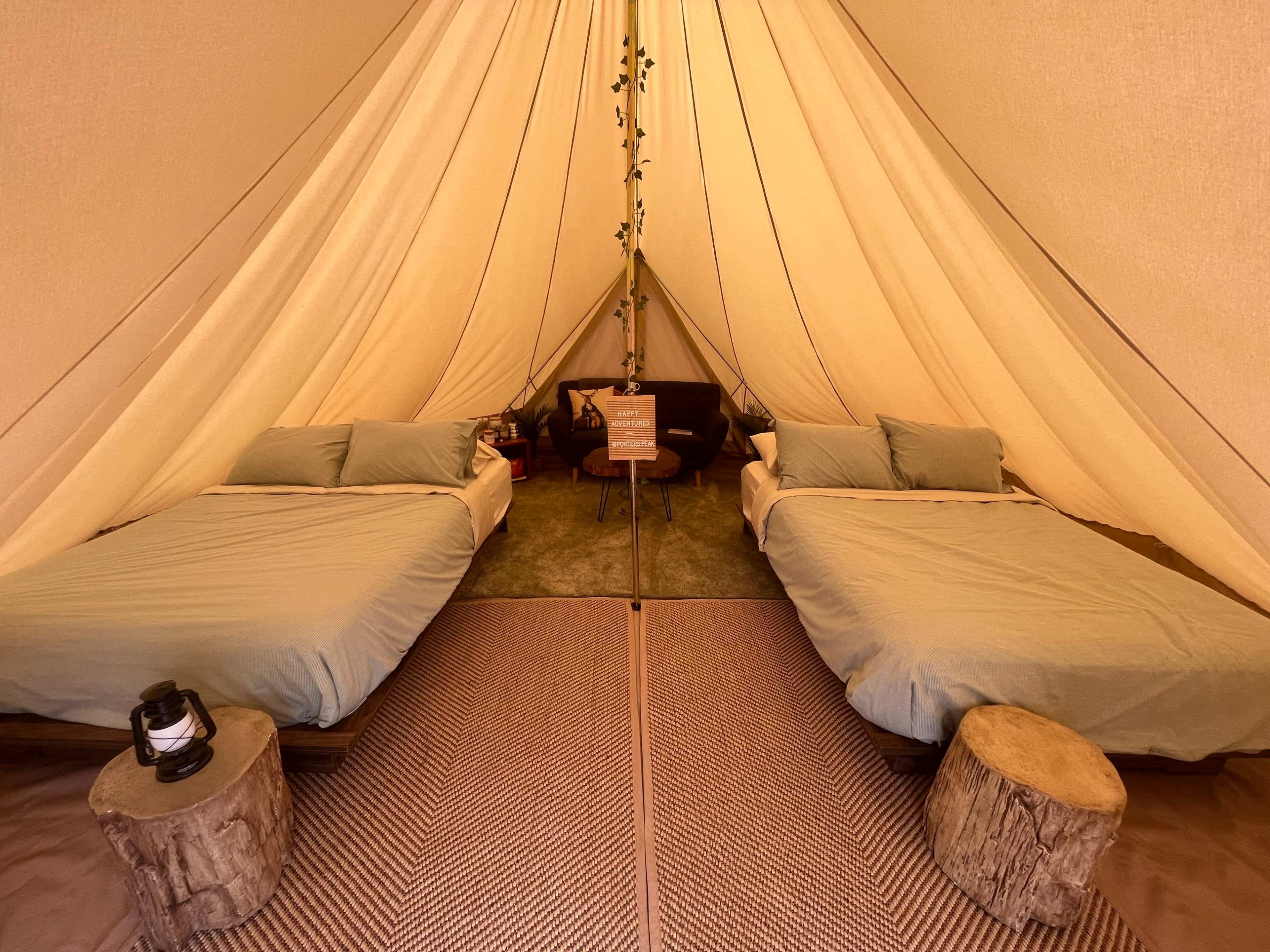 A spacious tent interior with two neatly made beds, a small table in the center, and natural wooden stools for seating.