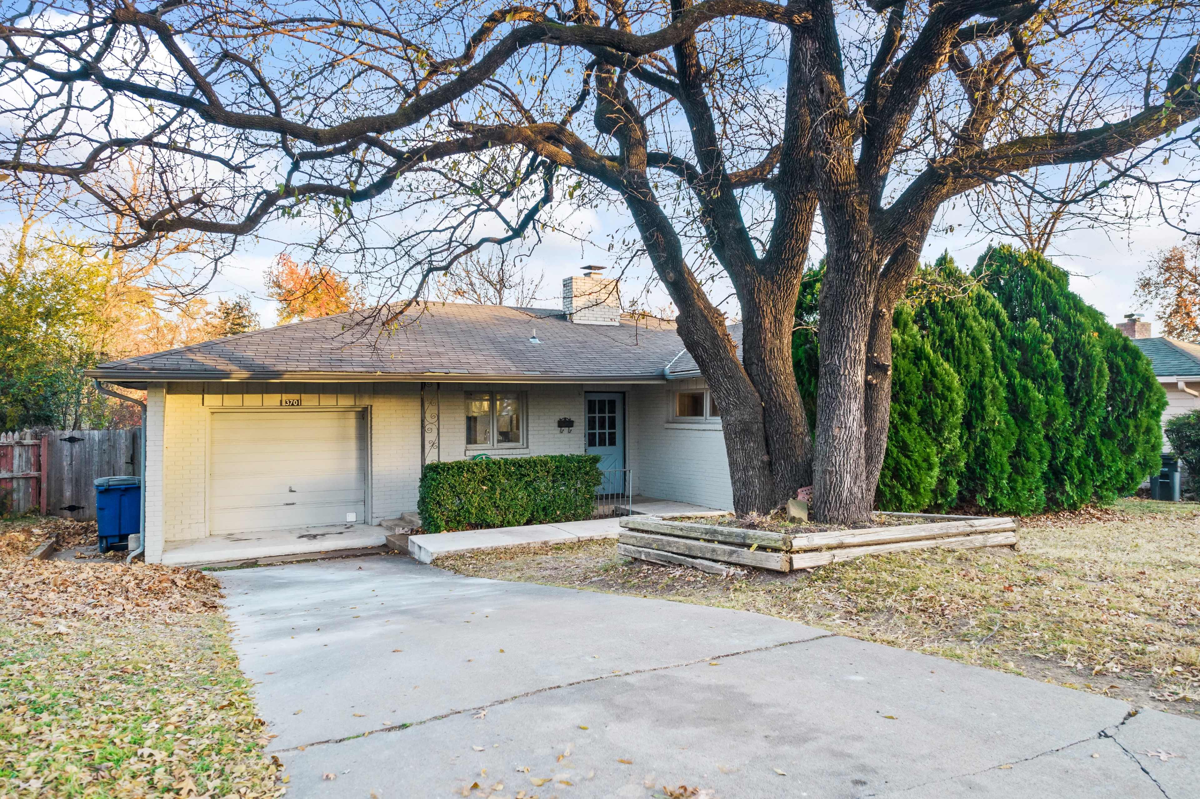 A single-story house with a garage is set back from a driveway lined by a large tree and evergreen shrubs.