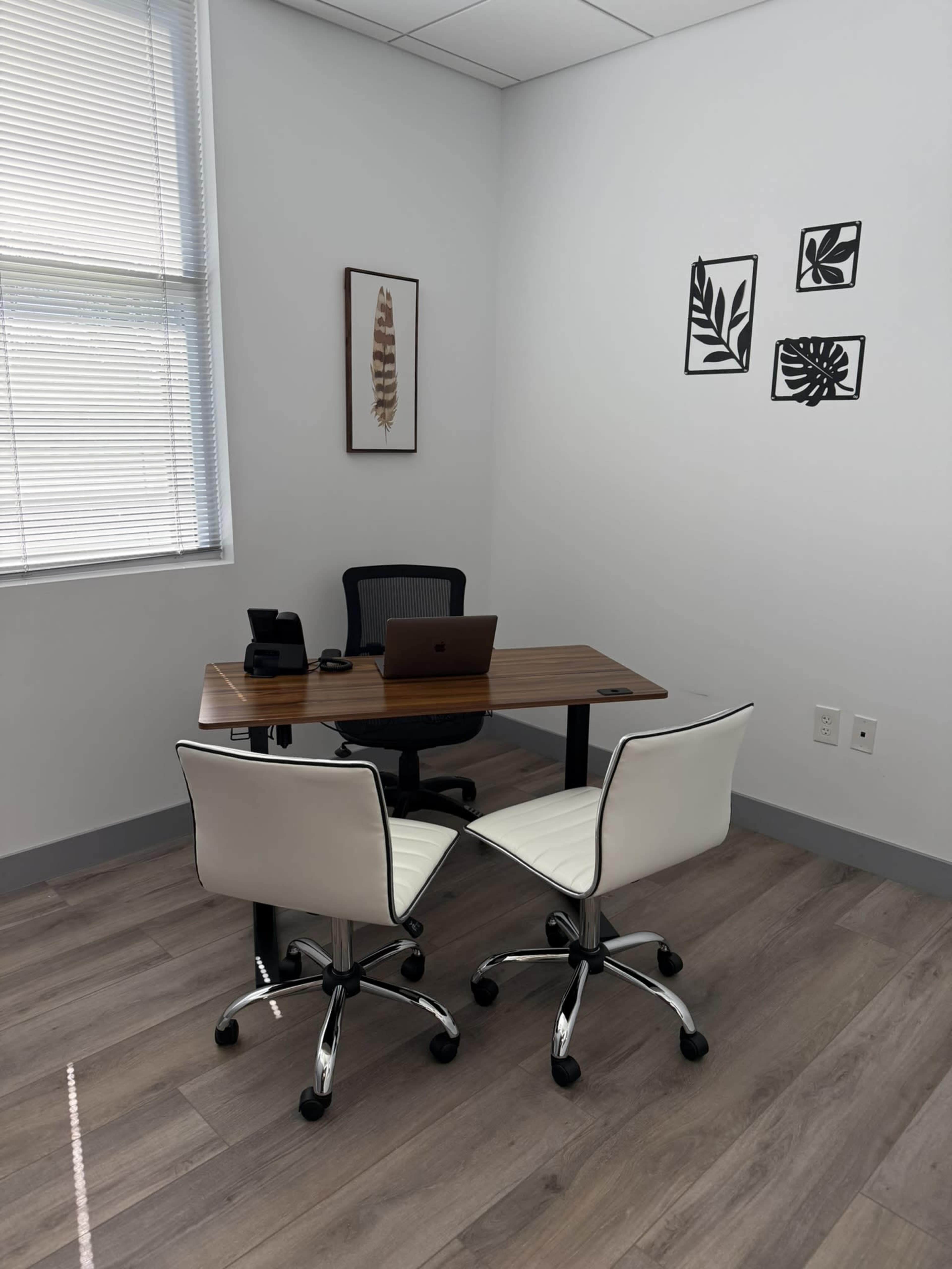 A small office features a wooden desk with a laptop and two white rolling chairs, situated against a wall adorned with framed leaf art.