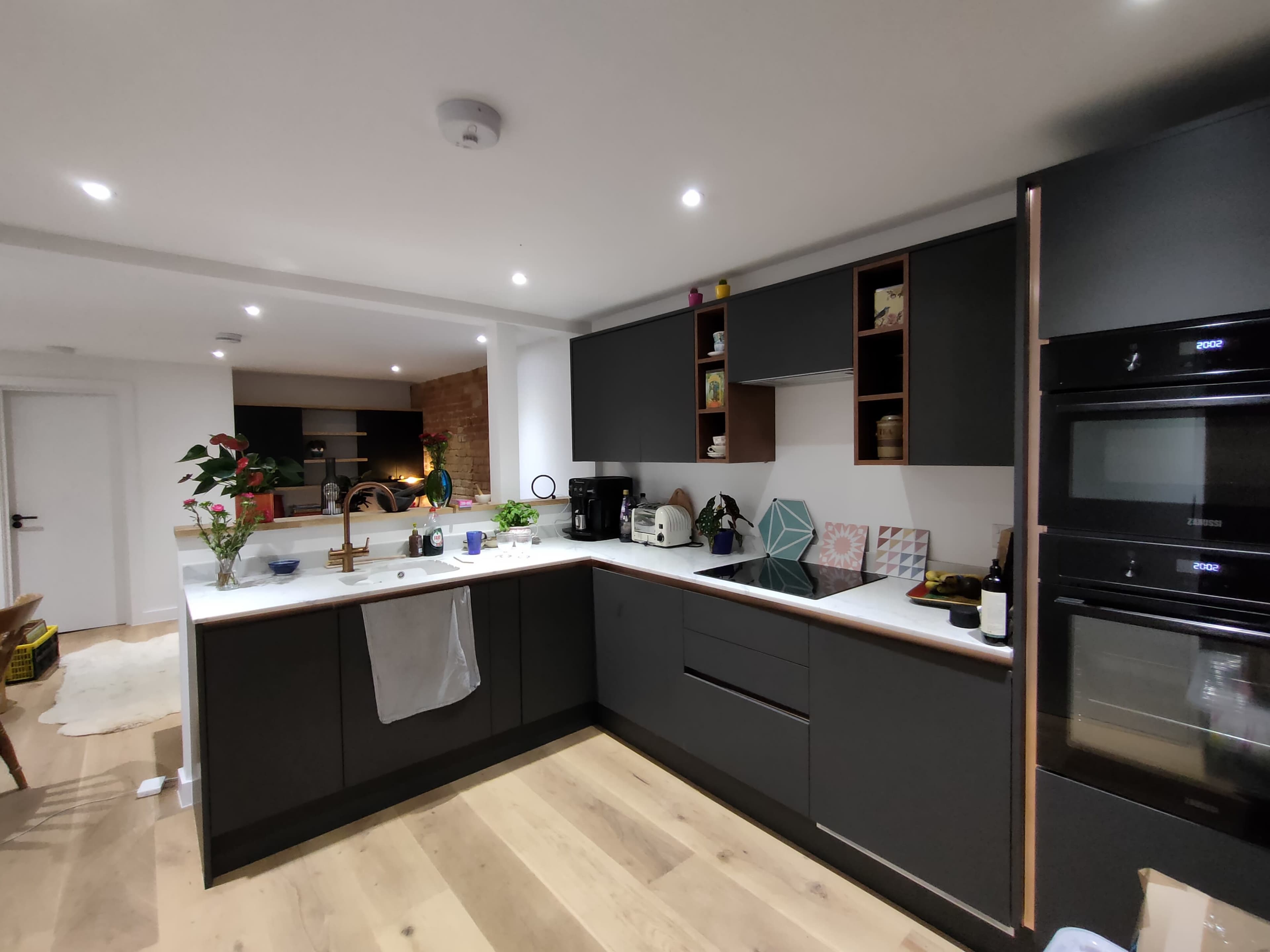 A modern kitchen with dark cabinetry, a white countertop, and various appliances, including an oven and coffee maker, alongside decor items on display.