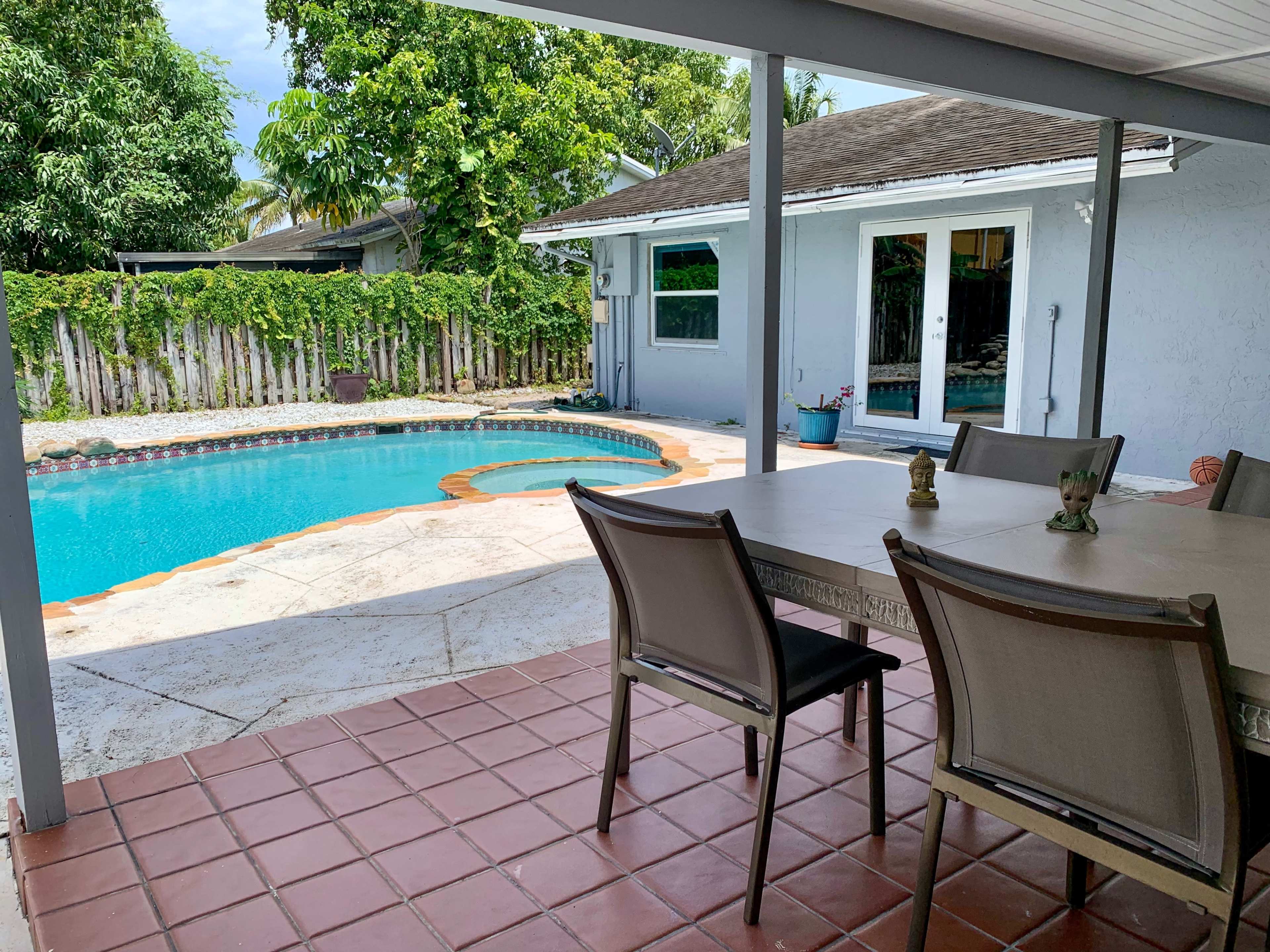 A patio area with a table and chairs overlooks a blue swimming pool surrounded by greenery and a house in the background.