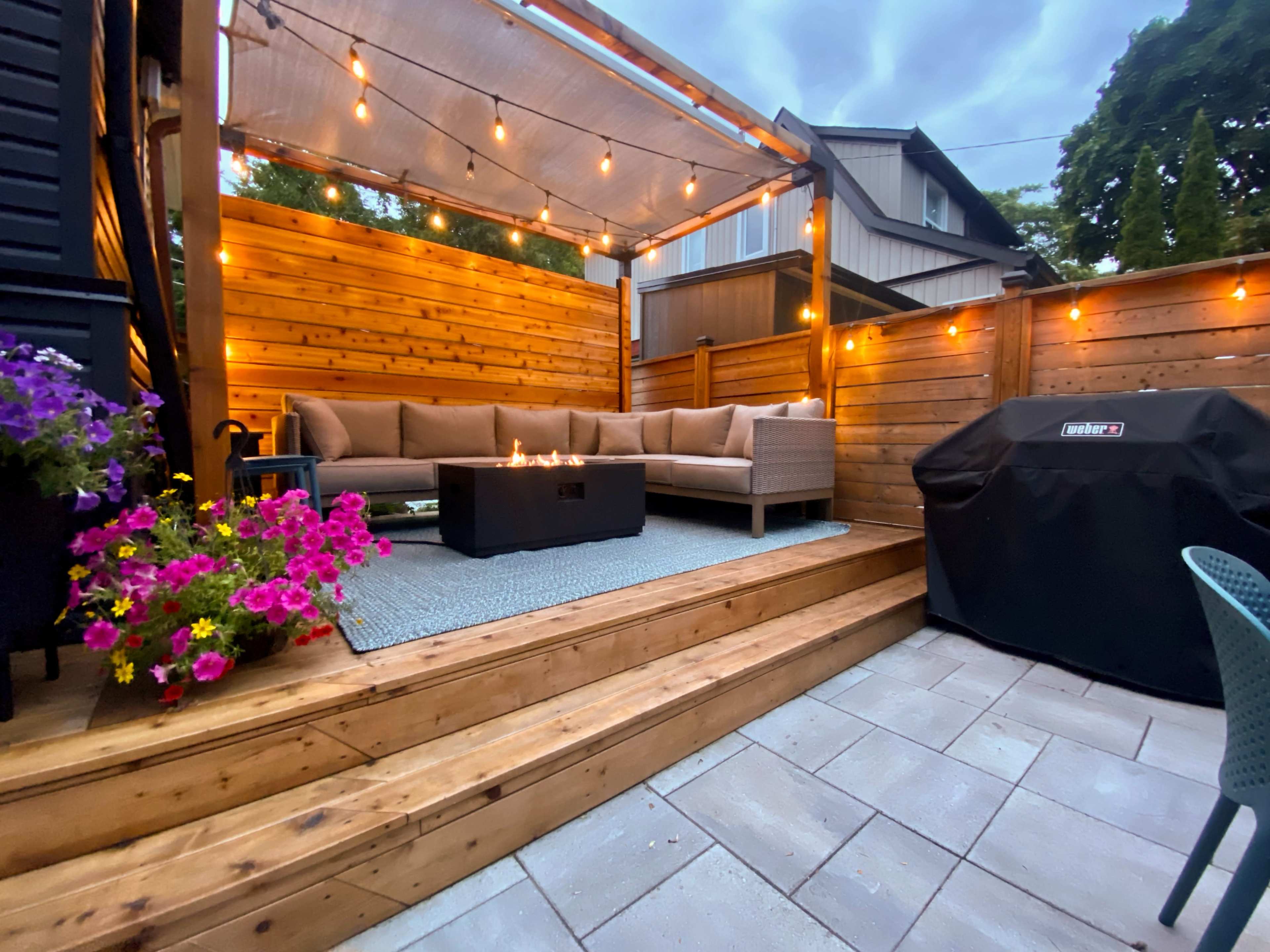 A wooden patio features a sectional sofa with a fire table, surrounded by string lights and potted flowers, alongside a grill in a well-lit backyard.
