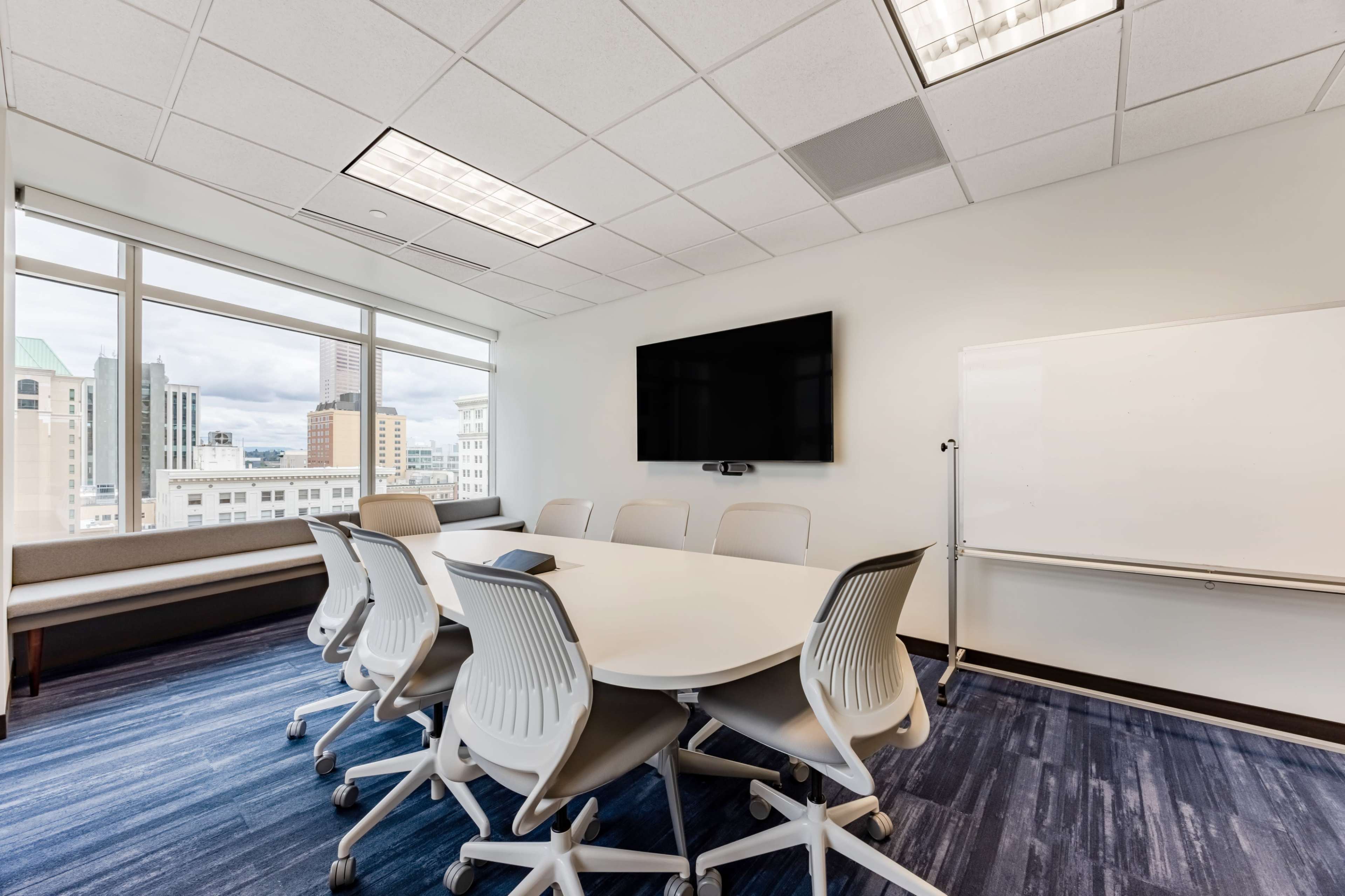 The image shows a modern conference room with a large round table, ergonomic chairs, a flat-screen TV mounted on the wall, and a whiteboard nearby, illuminated by natural light from large windows.