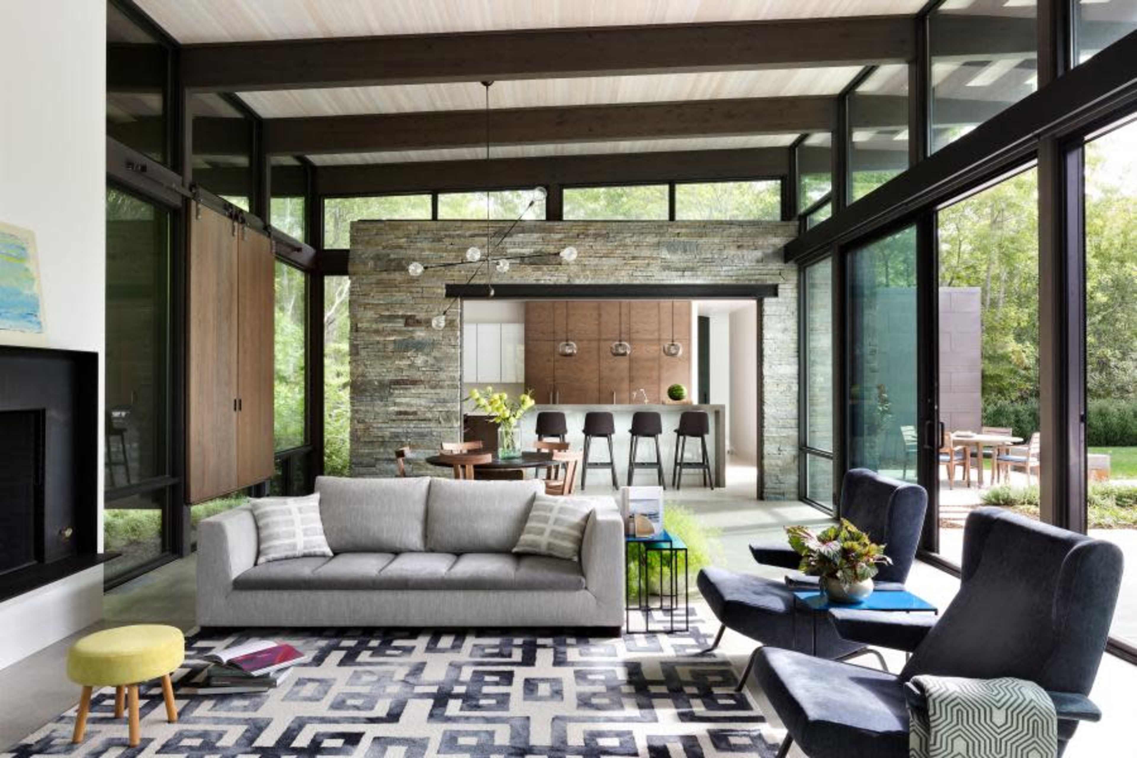 A modern living space with large windows, a gray sofa, and a view of an open kitchen and outdoor area.